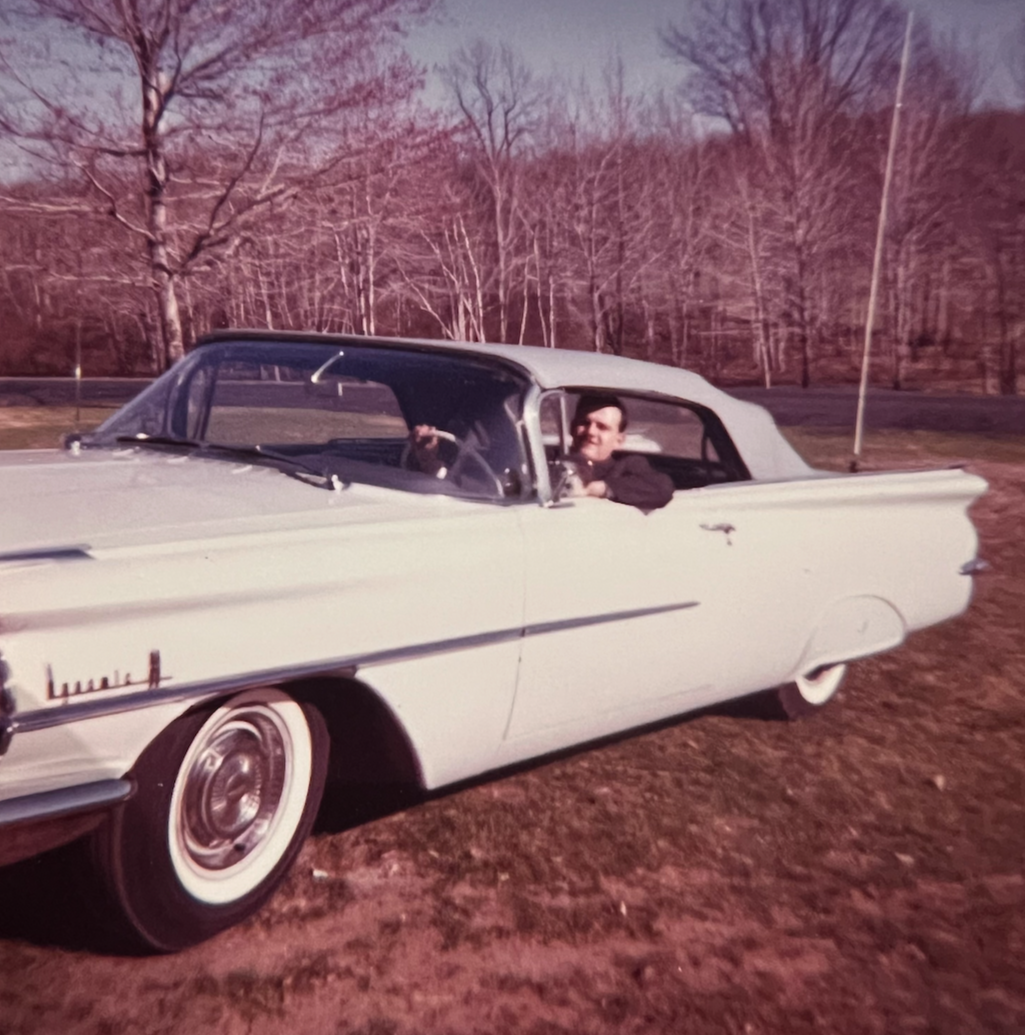 A vintage white convertible car parked on a grassy area with a man sitting in the driver's seat, holding the steering wheel, and trees in the background.