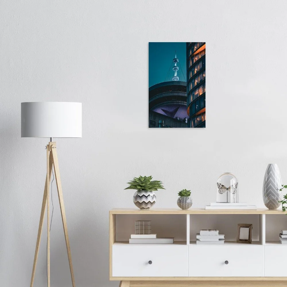 A modern living room with a white wall, a wooden floor lamp, a wooden and white bookshelf, and a framed cityscape photograph on the wall. The bookshelf has potted plants, decorative objects, and books.