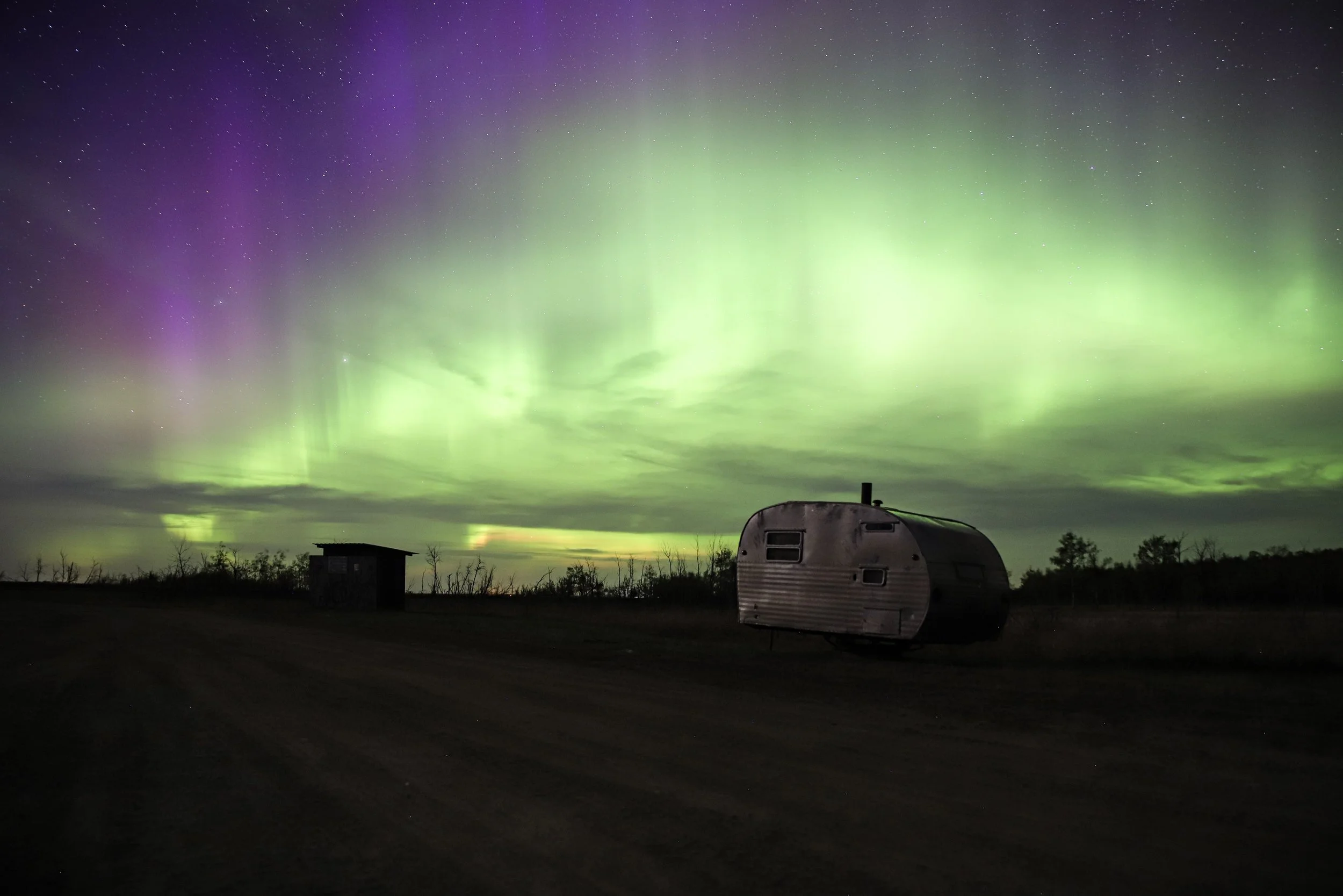 A dark landscape featuring a vintage camper trailer parked on a dirt path, with a small shed nearby, under a vibrant display of green and purple Northern Lights in the night sky. Taken by Paul Rophe In Alberta