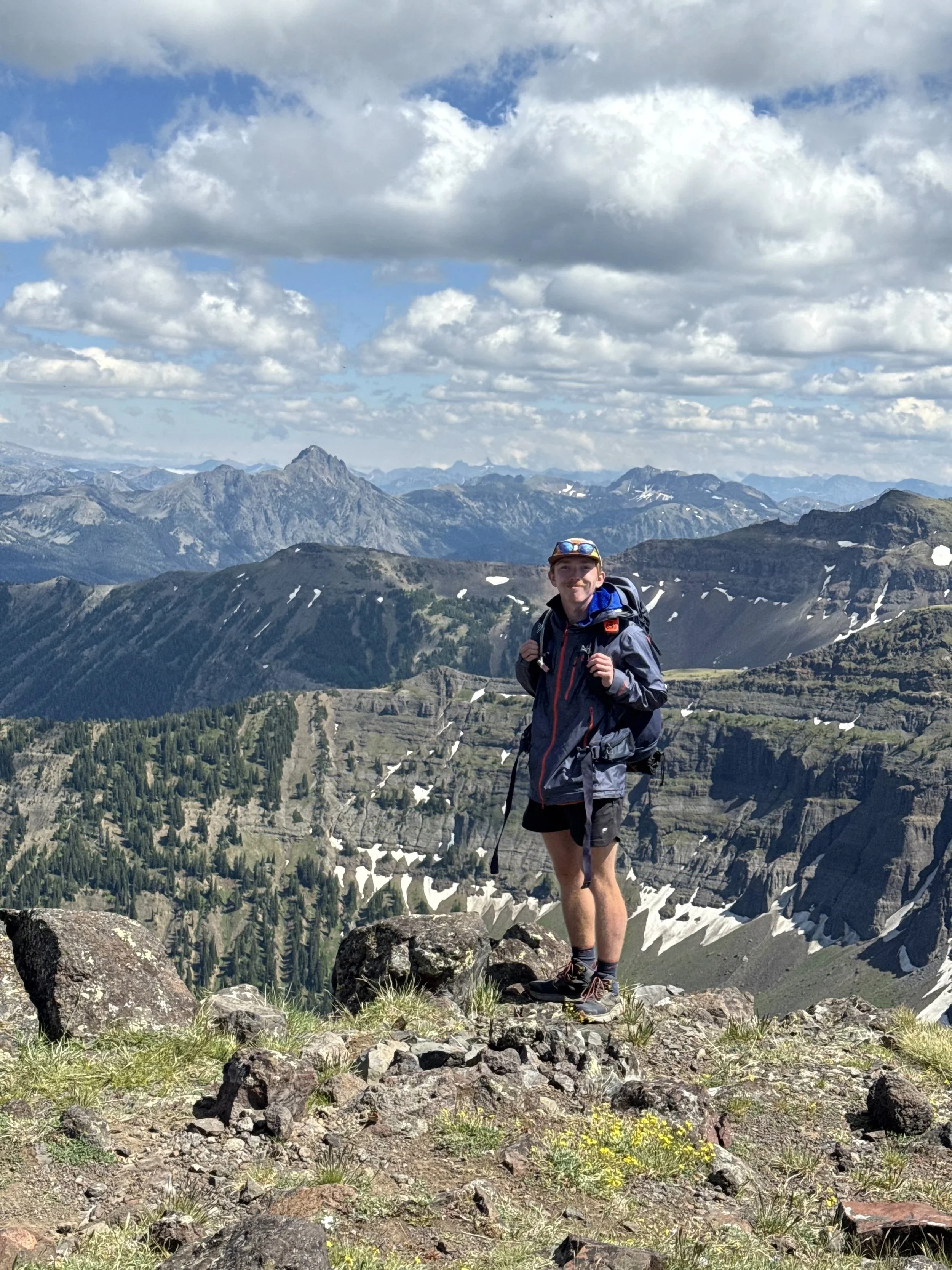A man in hiking gear standing on rocky terrain with mountains and cloudy sky in the background.