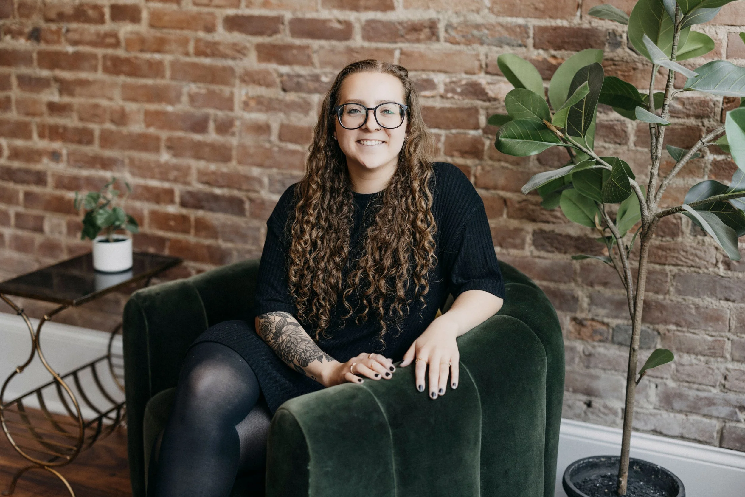 A young woman with long curly hair, wearing glasses and a black dress, sitting on a green velvet armchair in a room with a brick wall, next to a large green potted plant and a side table with a small potted plant.