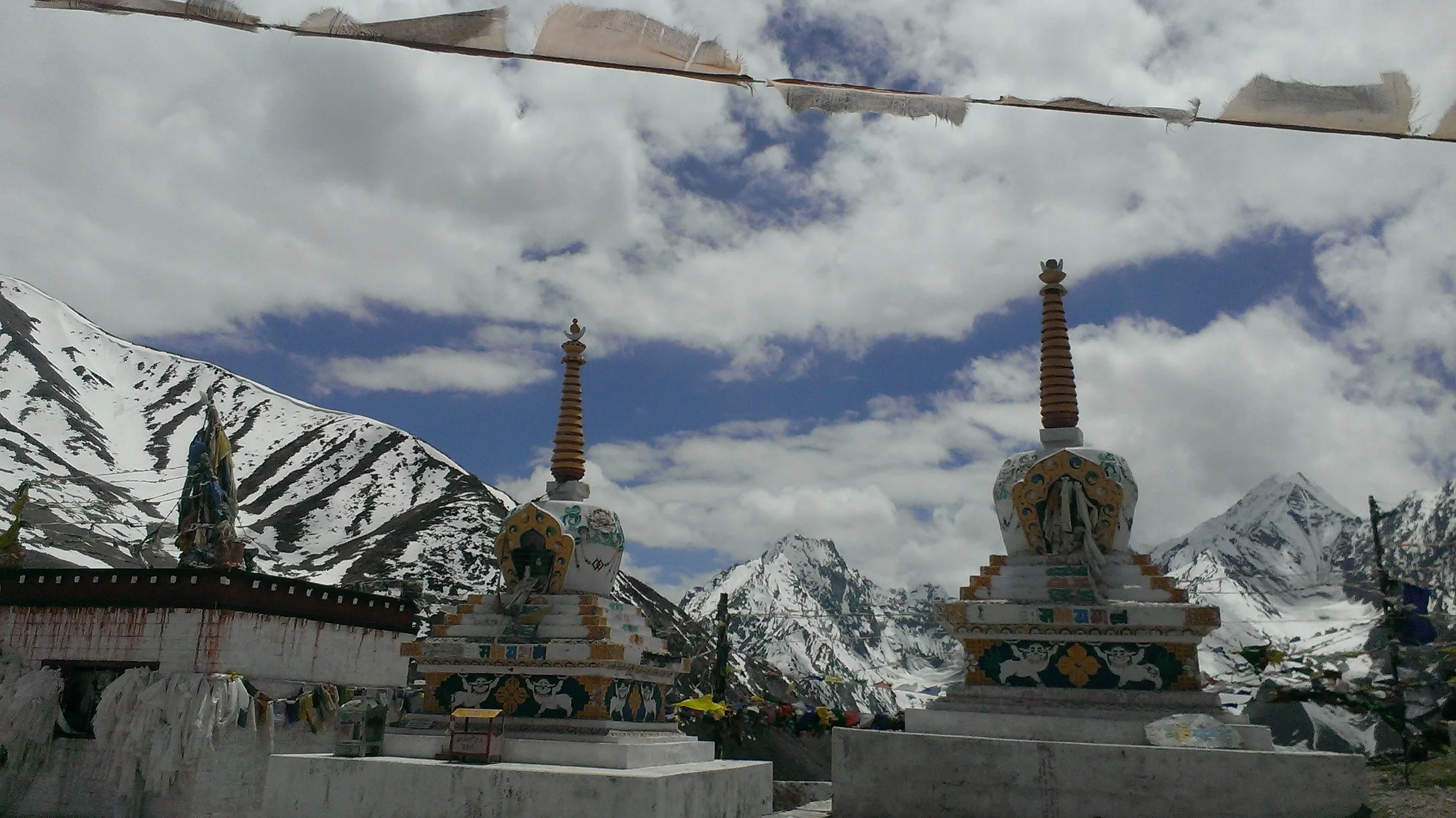 Vue de deux stupas colorés avec montagnes enneigées en arrière-plan, ciel partiellement nuageux, drapeaux de prière tibétains suspendus au sommet.