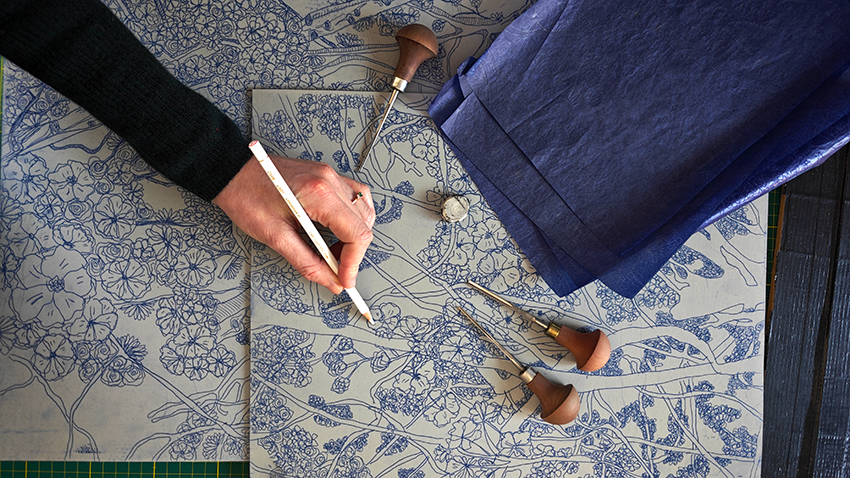 Une personne utilise un stylet sur un tissu de coton avec un motif de broderie, entourée de divers outils de couture, dont des épingles et un dé à coudre.