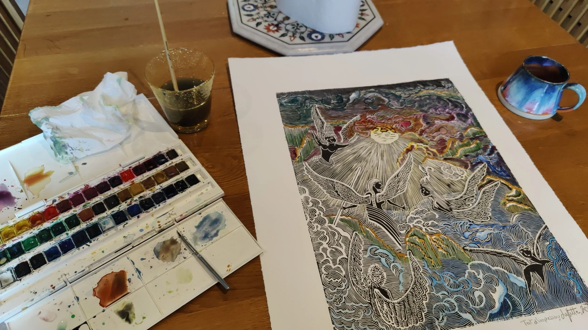 Une table en bois avec une palette de peinture aquarelle, un pinceau, un verre de boisson, un moule avec un papier peint avec une illustration colorée d'anges ou de figures mythologiques, une tasse colorée, et un mouchoir ou essuie-tout. Le tableau semble être une œuvre d'art en cours de création.