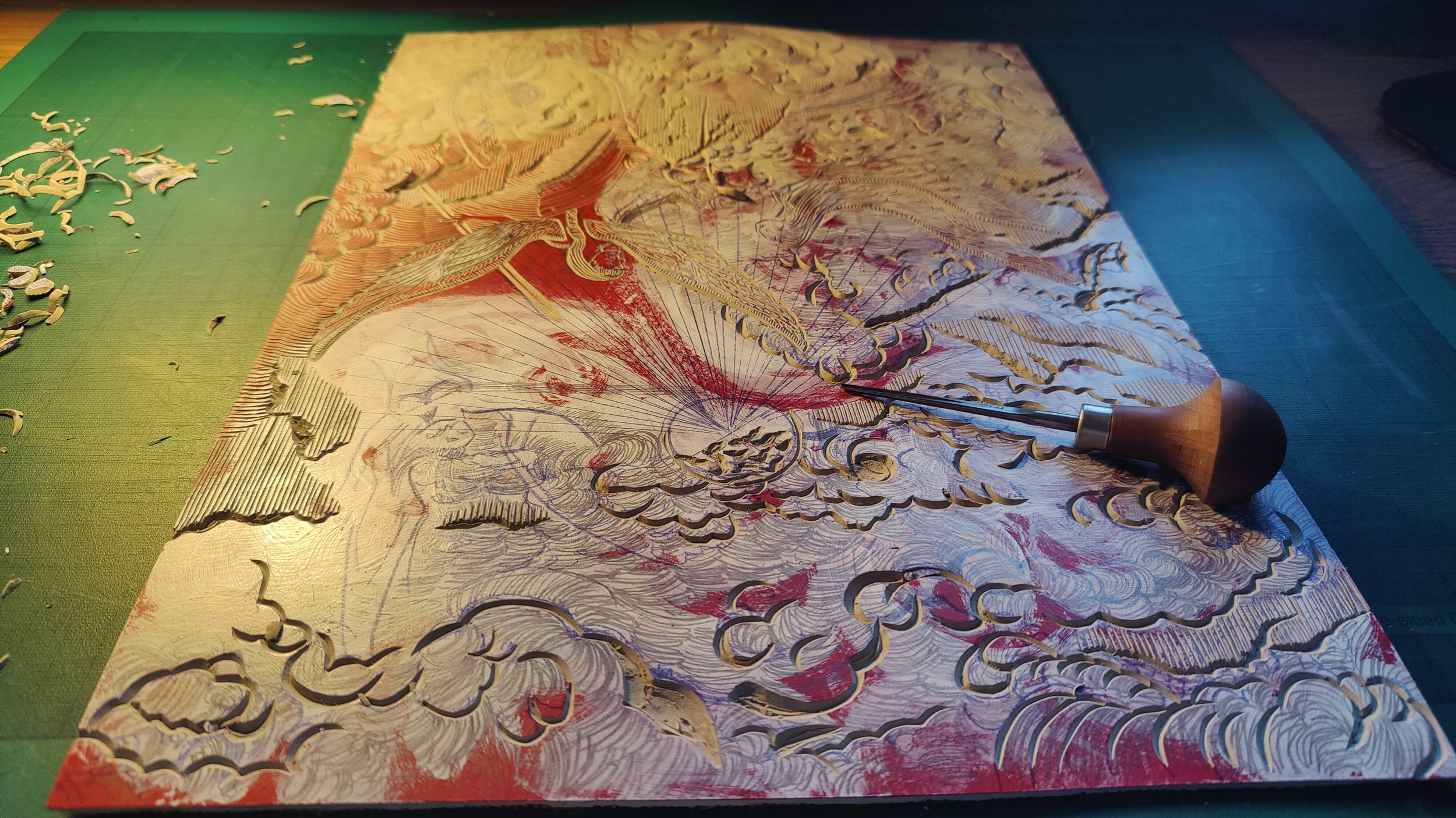 Une plaque gravée en cours de création avec un burin, représentant une scène avec des nuages, des oiseaux et un soleil, sur une surface en bois colorée.