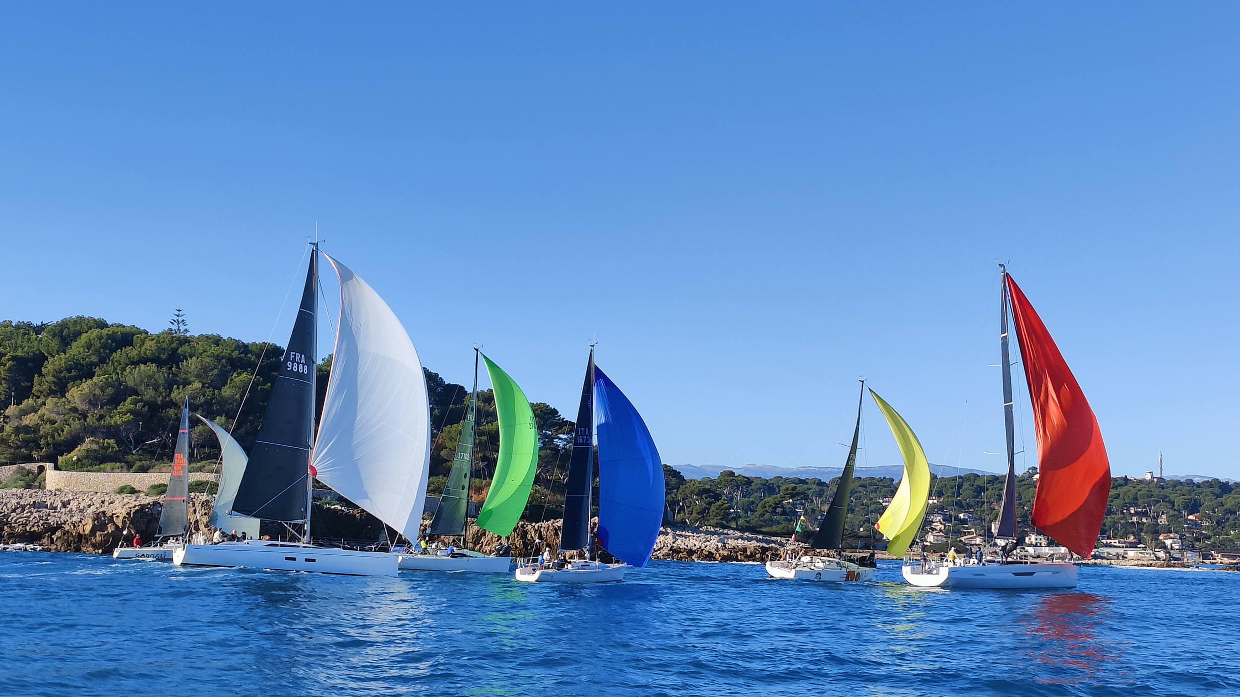 Bateaux de course à voile avec voiles colorées, en mer, avec un littoral verdoyant en arrière-plan sous un ciel clair.