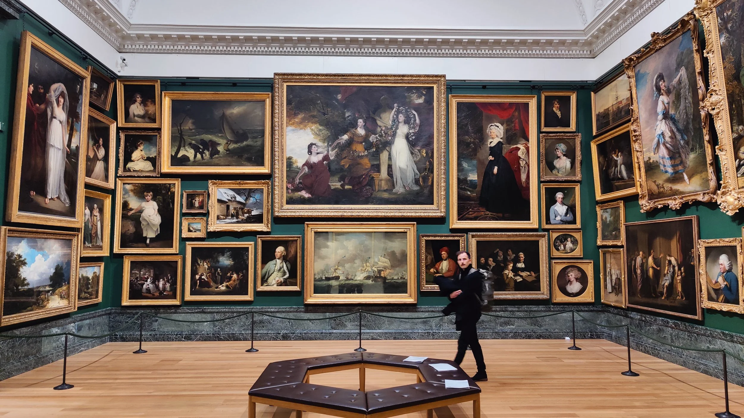 Une salle d'exposition avec de nombreux tableaux classés dans des cadres dorés sur les murs, un banc au centre, un jeune homme avec un sac à dos selles devant les œuvres.
