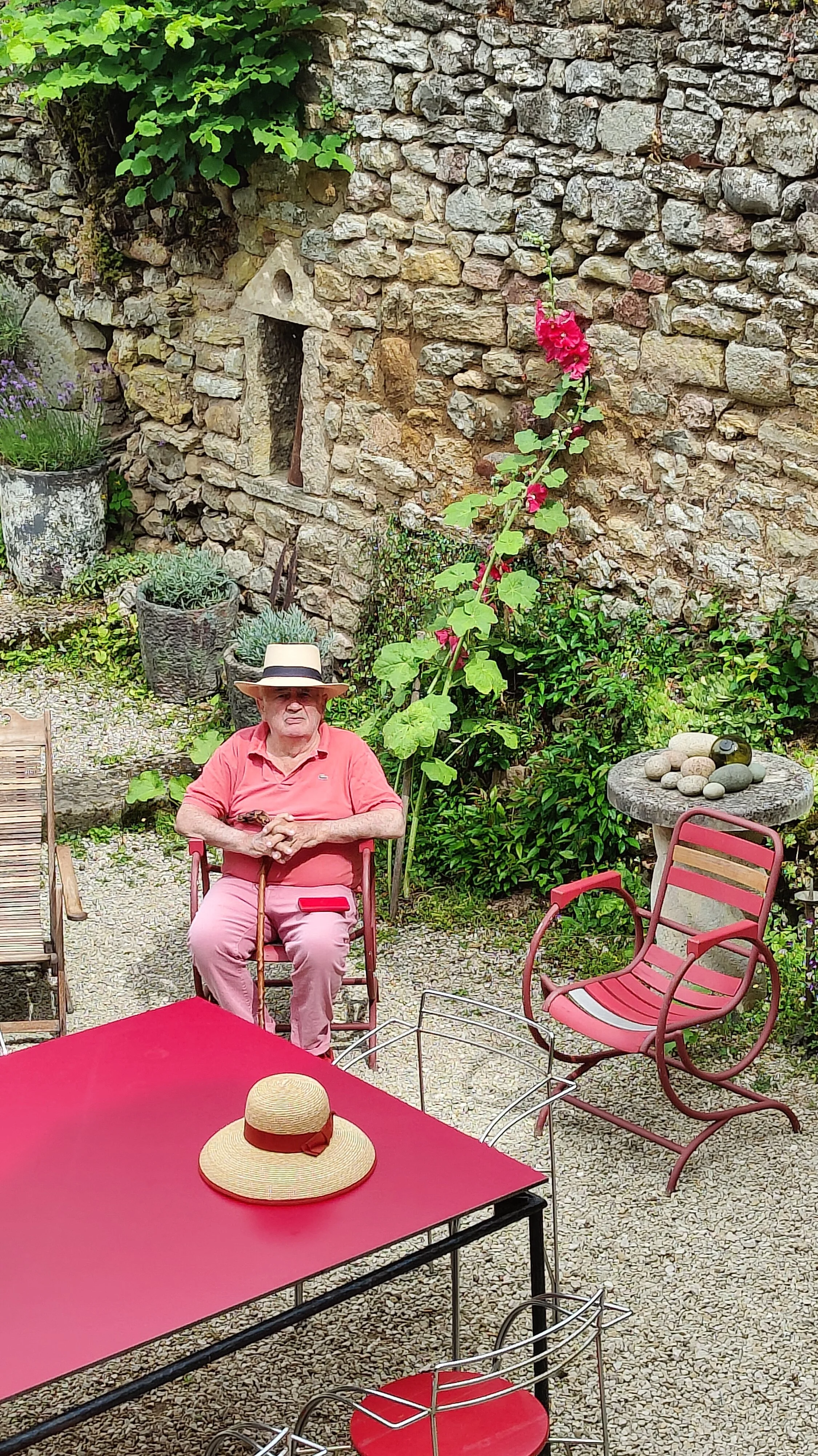 Un homme âgé portant un chapeau, assis sur une chaise dans un jardin avec une table rouge, un chapeau de soleil posé dessus, et entouré de mobilier de jardin. Le fond montre un mur en pierre avec des plantes et des fleurs, notamment une grande tige d