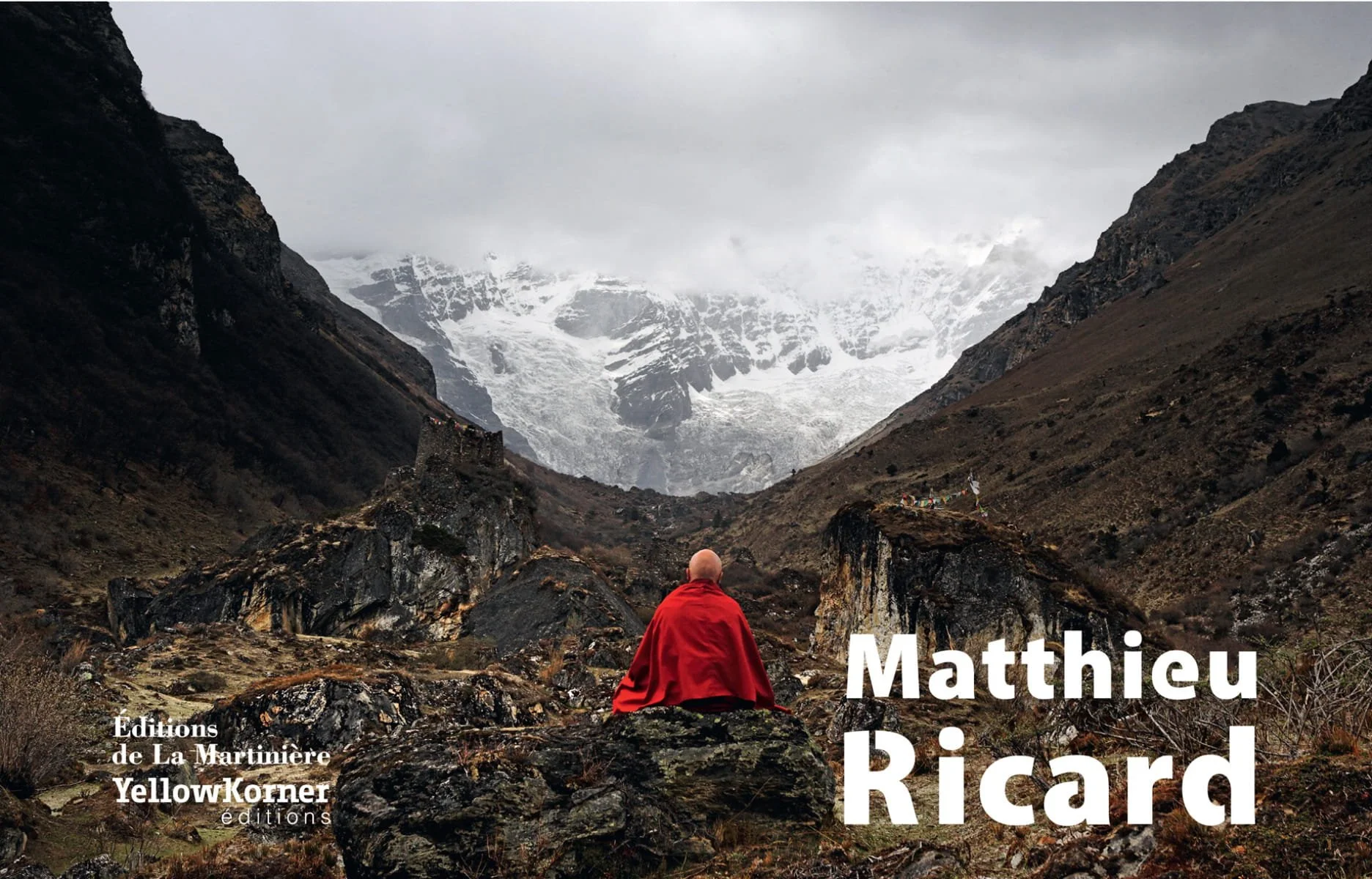 Un moine en robe rouge assis dans une vallée montagneuse, entouré de rochers et de montagnes enneigées en arrière-plan.