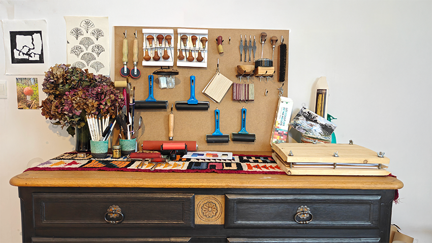 Un atelier d'artisan avec un bureau en bois foncé, une planche à outil en liège avec divers outils de sculpture, pinceaux, rouleaux, et fiches, ainsi que des papiers et un bouquet de fleurs dried sur le bureau.