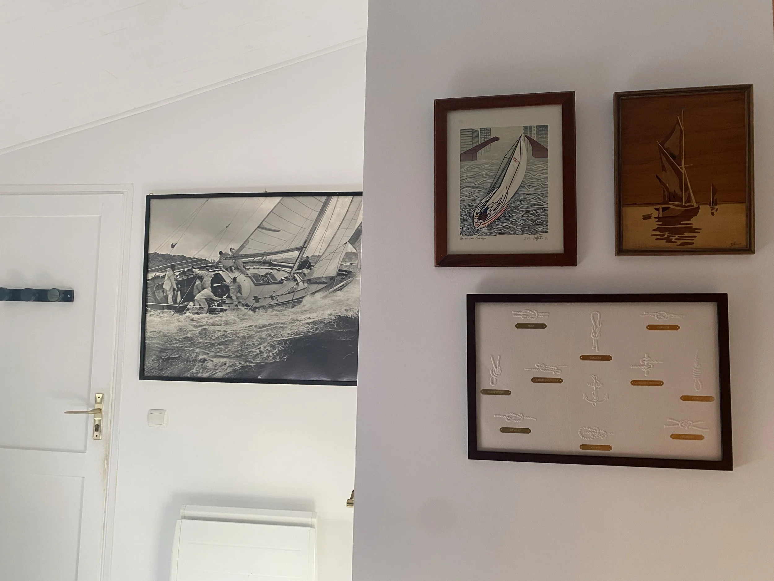 Mur blanc décoré avec un tableau noir et un tableau blanc encadrés d'environ 30 cm, affichant des illustrations de voiliers, accrochés à une surface verticale dans une pièce. À gauche, un grand tableau en noir et blanc représentant une scène de navigation à la voile avec des personnes sur un bateau.