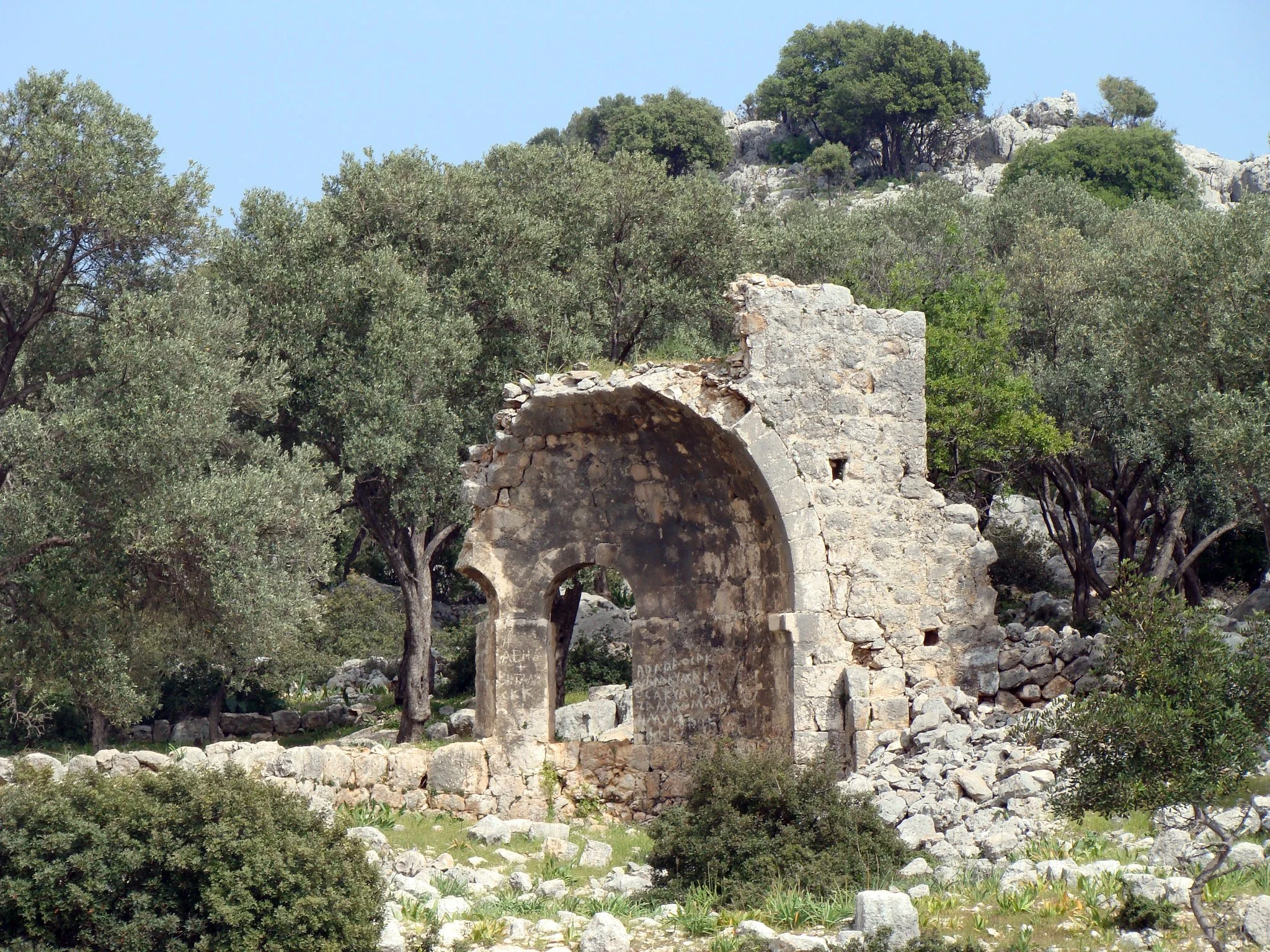 Restes d'une ancienne structure en pierre, probablement une porte ou un arc, entourée de végétation et d'arbres, sous un ciel clair.