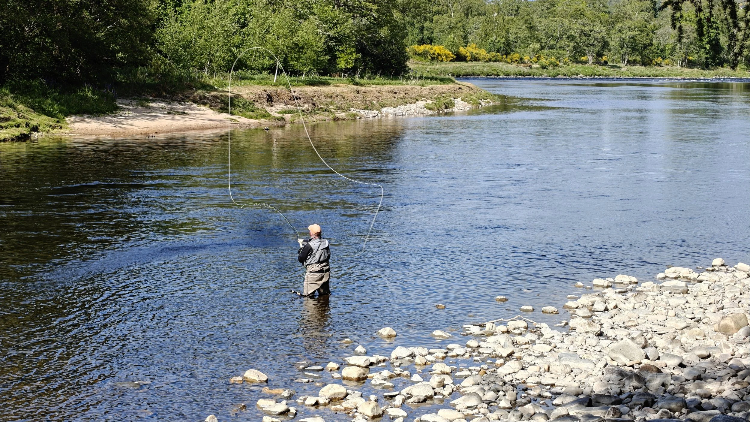 Un pêcheur en waders pêche dans une rivière calme entourée de verdure et de rochers sur le rivage.