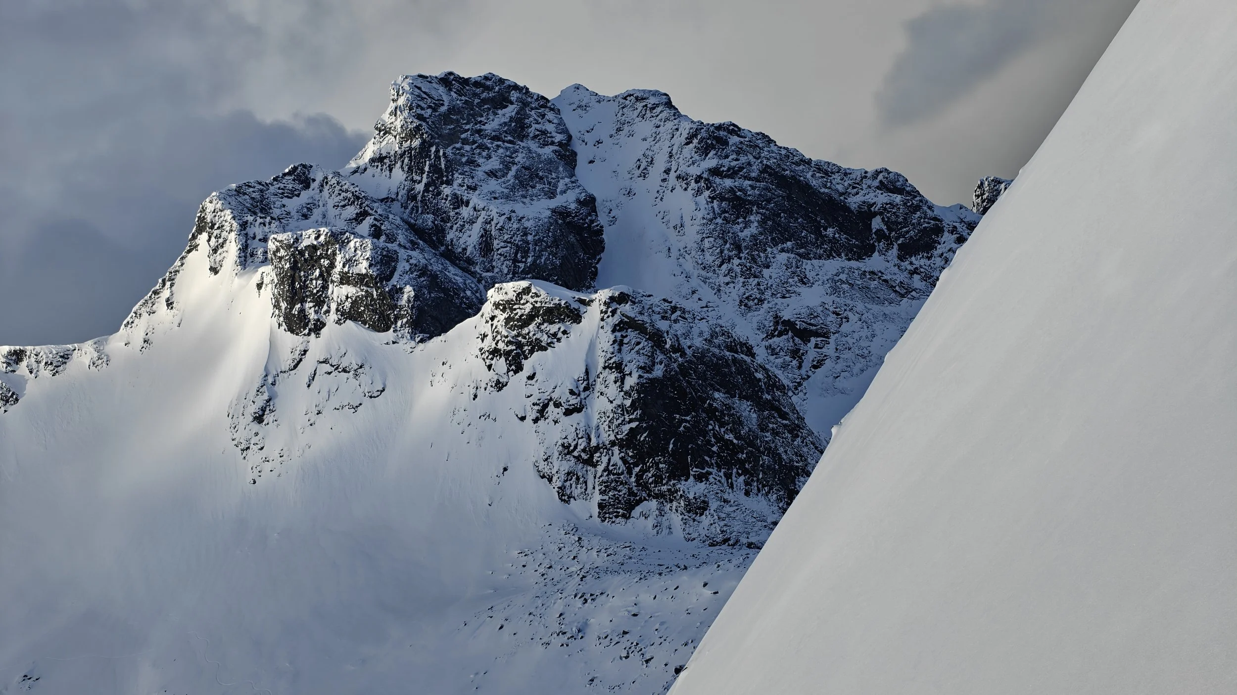 Montagnes enneigées avec un ciel nuageux, vue d'une pente enneigée sur le côté droit de l'image.