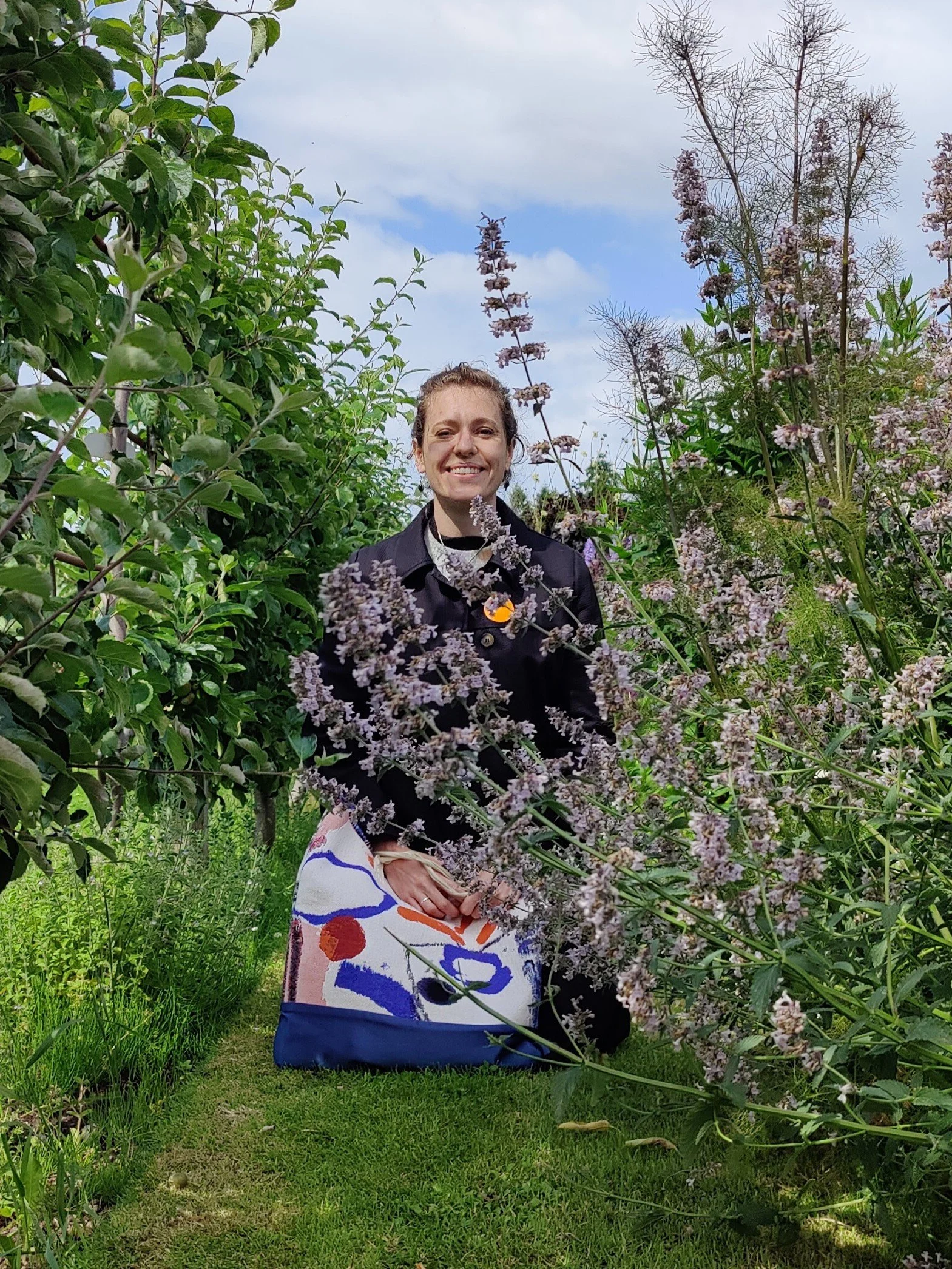 Une femme souriante se trouve au milieu de buissons de fleurs violettes, avec un ciel partiellement nuageux en arrière-plan.