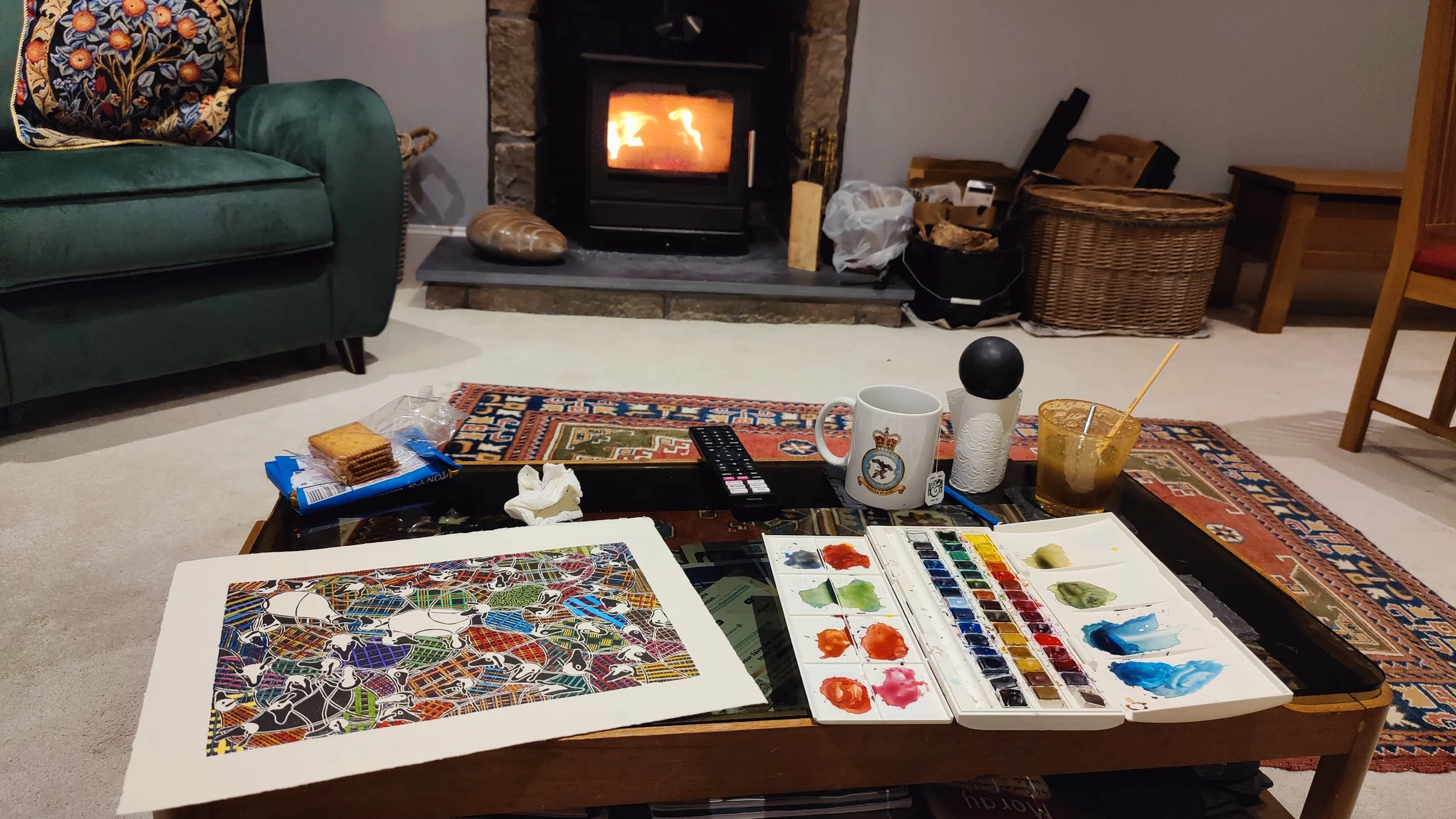 Un salon avec une cheminée allumée, un tapis, un canapé vert, une table basse avec de la peinture, des pinceaux, une tasse, un verre à boule, et des biscuits, dans une ambiance chaleureuse