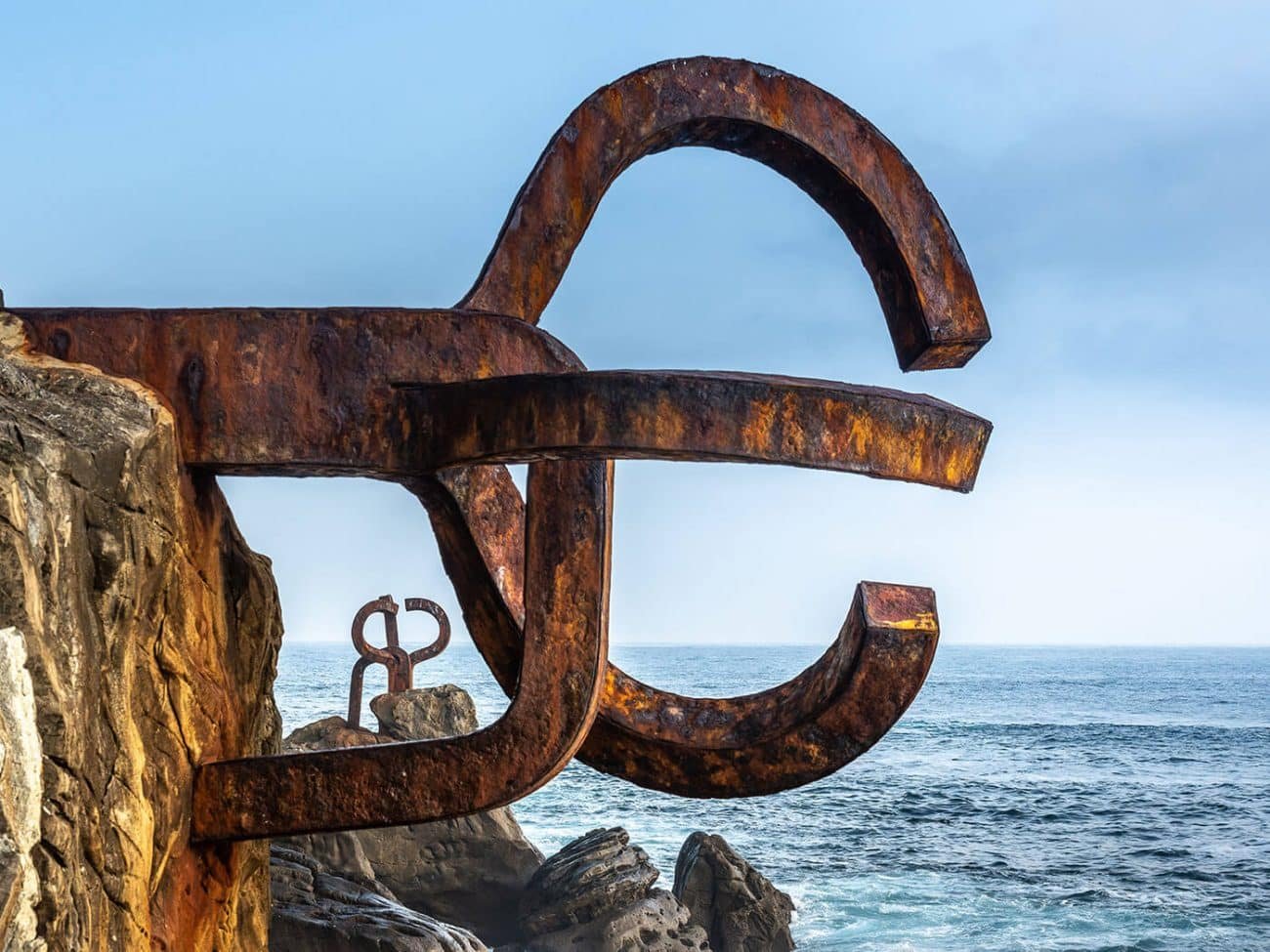 Sculpture en métal rouillé représentant un symbole euro, située sur une falaise avec vue sur la mer.