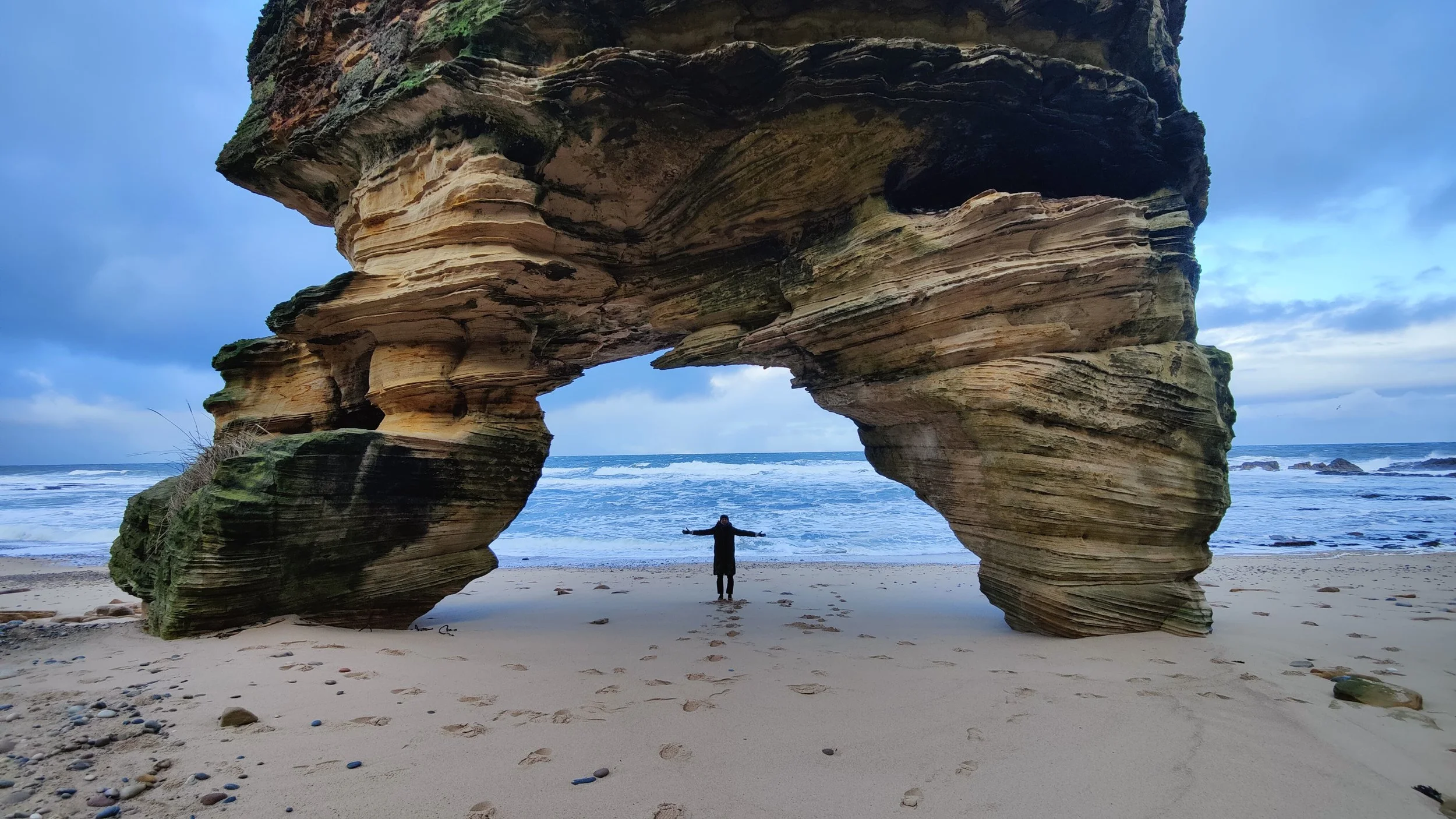 Une personne se tenant avec les bras ouverts sous un grand rocher en forme d'arche sur une plage, avec la mer en arrière-plan et un ciel nuageux.