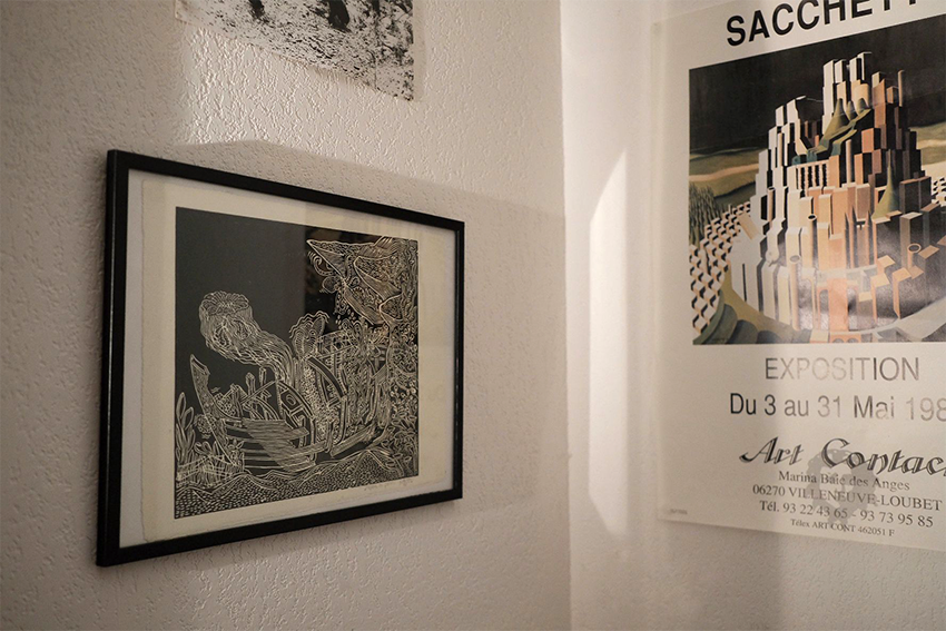 Photographie d'une pièce avec un cadre noir contenant une œuvre d'art en noir et blanc, et un poster promotionnel d'une exposition d'art, accroché à un mur blanc.