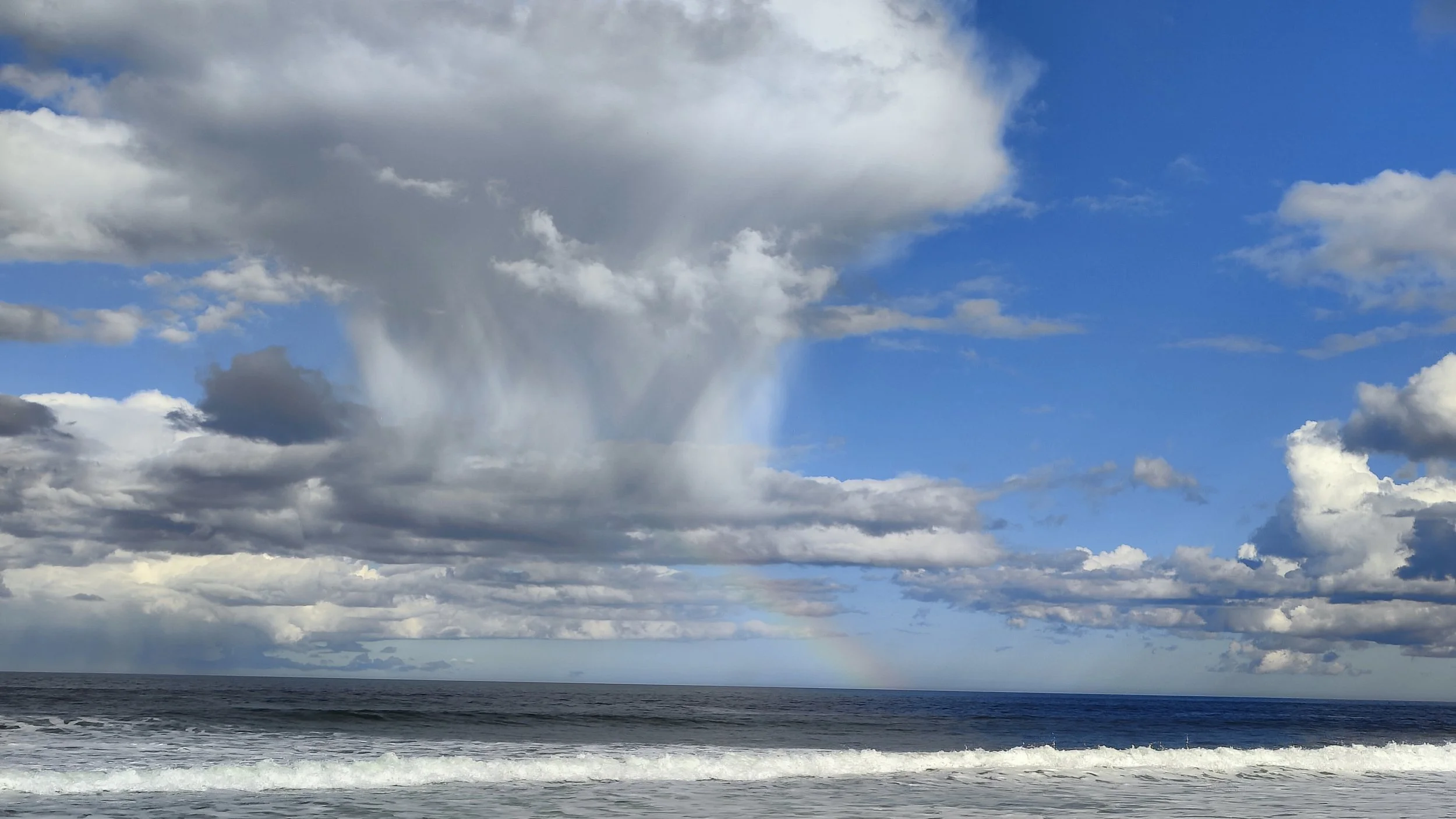 Ciel nuageux au-dessus de la mer, avec des vagues et un arc-en-ciel faint en arrière-plan.
