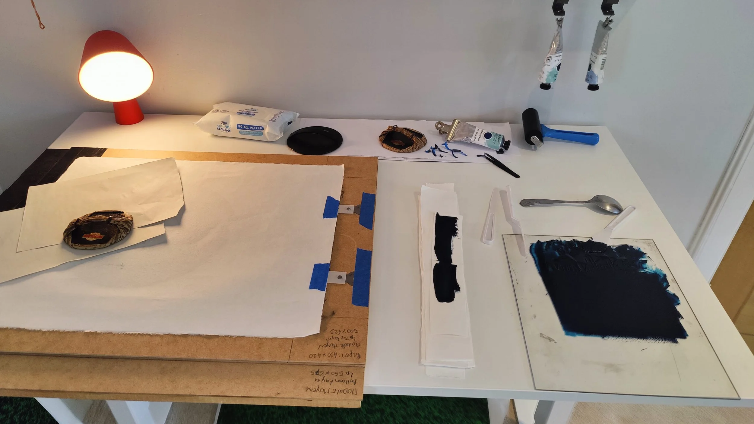 Surface de travail avec divers outils pour peinture ou artisanat, dont un rouleau en main, des pinceaux, de la peinture noire, un tissu, une lampe, du papier, et d'autres petits objets, sur un fond blanc et une table en bois.