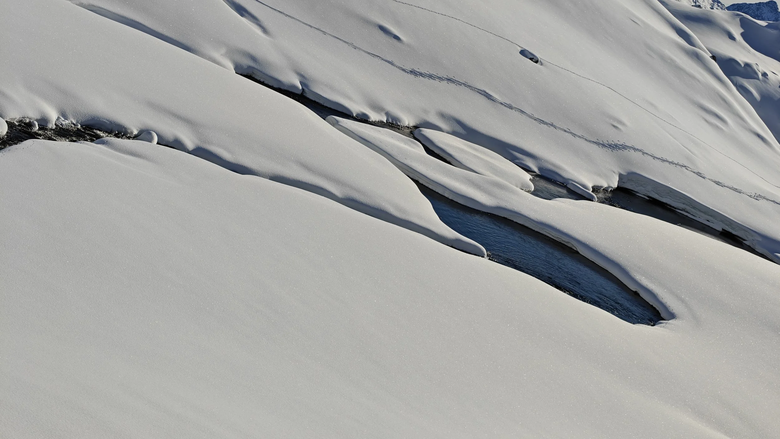 Neige couvrant partiellement un ruisseau gelé avec des rochers et de la glace visible.