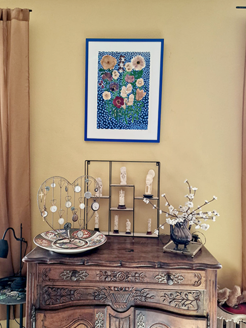 Une console ancienne en bois avec des décorations, des figurines en ivoire, un vase avec des branches blanches en fleurs, un cadre en métal noir avec des figurines en ivoire, une œuvre d'art florale dans un cadre bleu accroché au mur derrière.