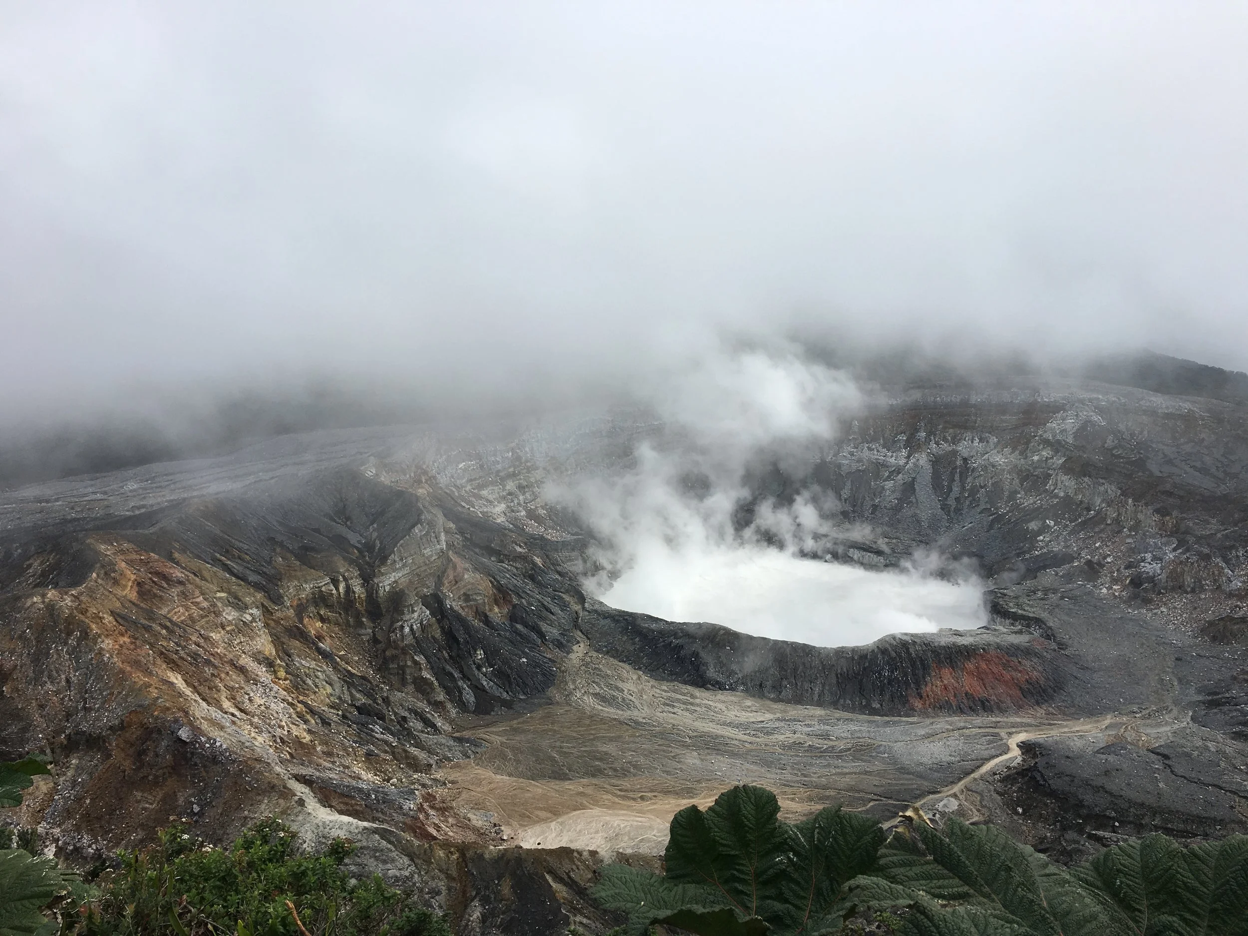 Vue d'un volcan en activité avec des fumées et des vapeurs s'échappant du cratère, entouré de roches et de végétation sparse, sous un ciel nuageux.