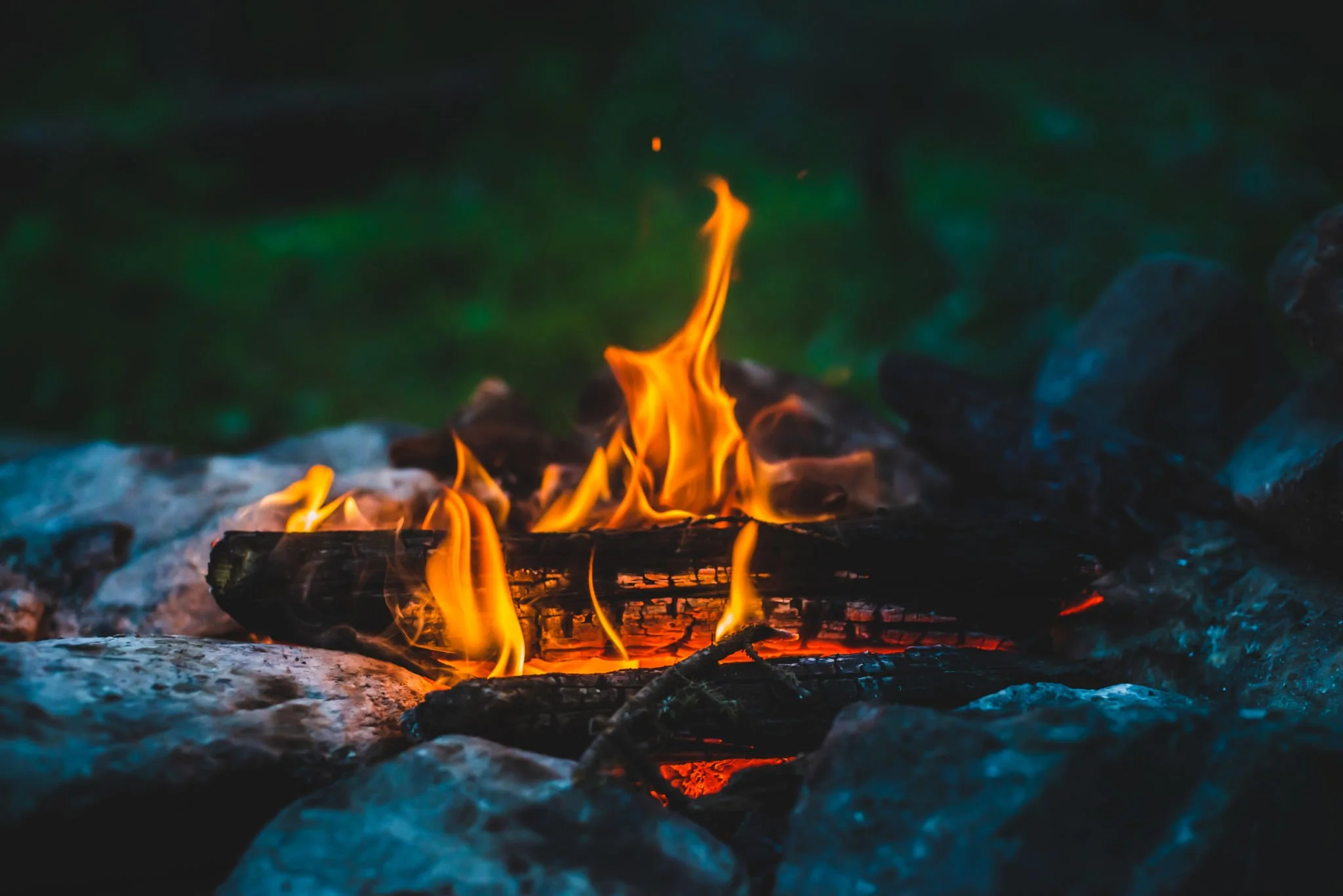 Close-up of small campfire with orange and yellow flames burning on a log, surrounded by rocks, with dark, blurred background.