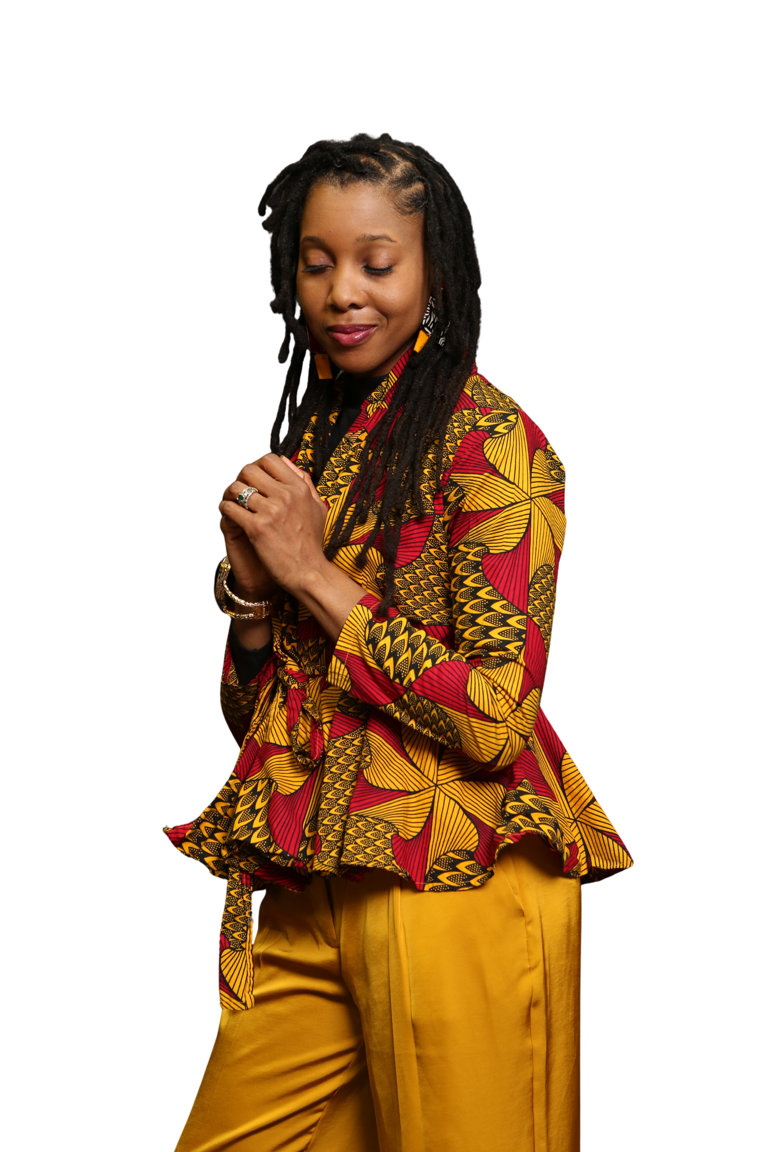 A woman with dreadlocks wearing a colorful patterned top and yellow pants, with her eyes closed and hands clasped.