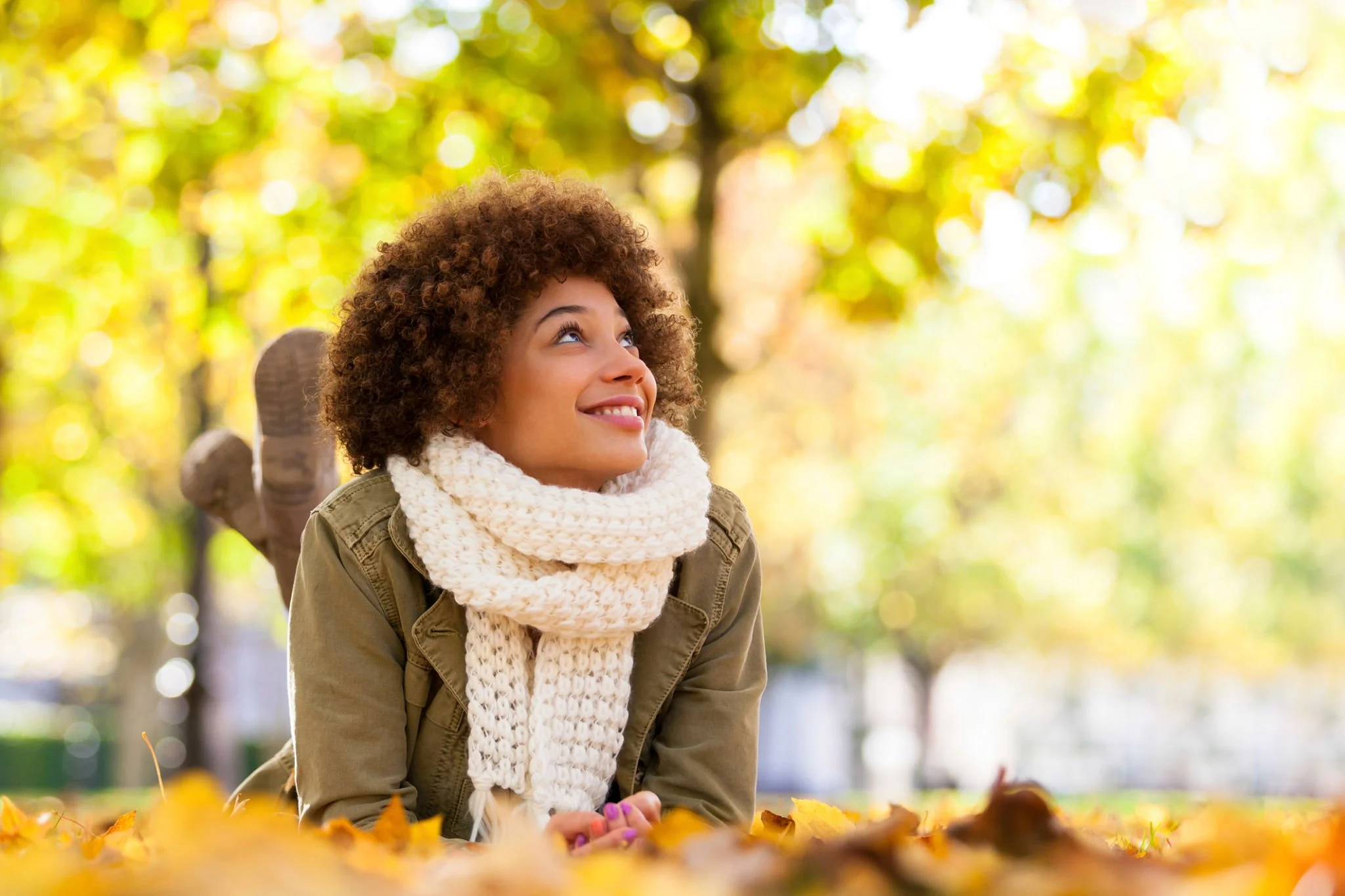 A young woman with curly hair, smiling and looking up, lying on the ground covered with autumn leaves in a park during fall, wearing a scarf and a jacket.