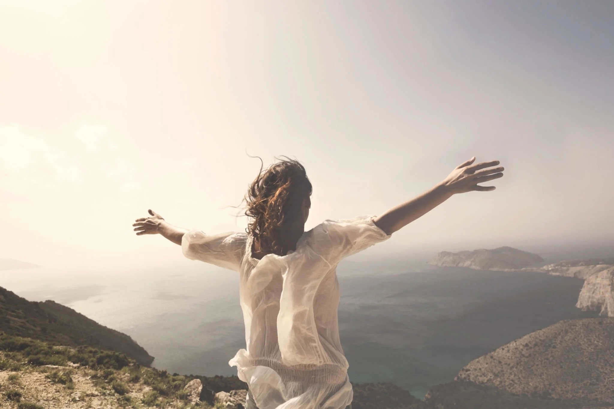 Woman with outstretched arms on a hilltop overlooking the ocean, wearing a light, flowing shirt, with mountains and water in the background under a bright sky.