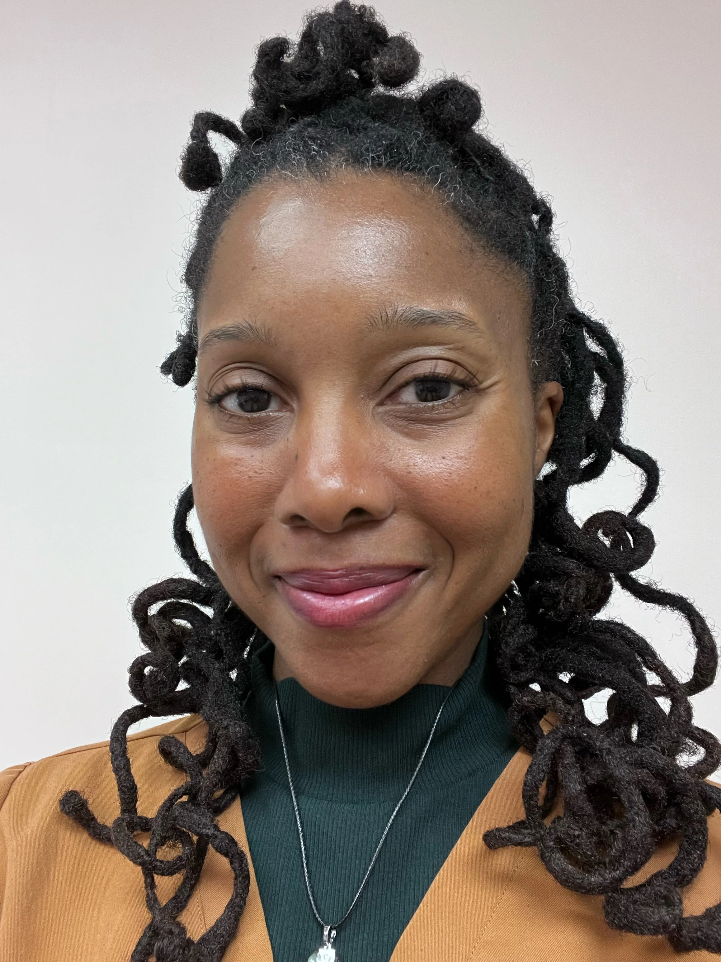 Close-up of a woman with dark, curly dreadlocks, smiling, wearing a green top, tan blazer, and a silver necklace with a pendant, against a plain light-colored background.
