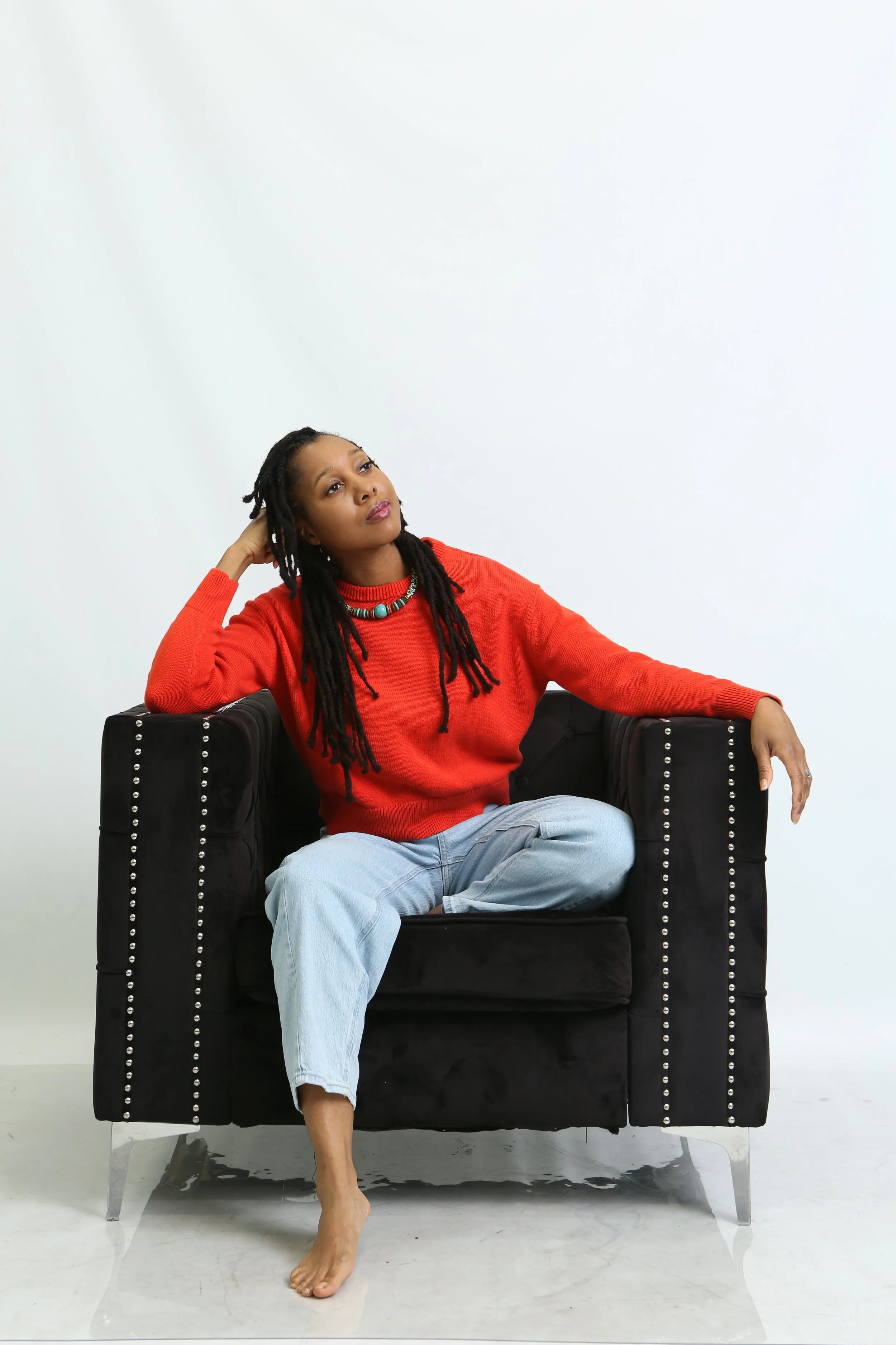 A woman with long dreadlocks sitting on a black armchair with silver nailhead trim, wearing a red sweater and light blue jeans, barefoot, with a thoughtful expression against a plain white background.