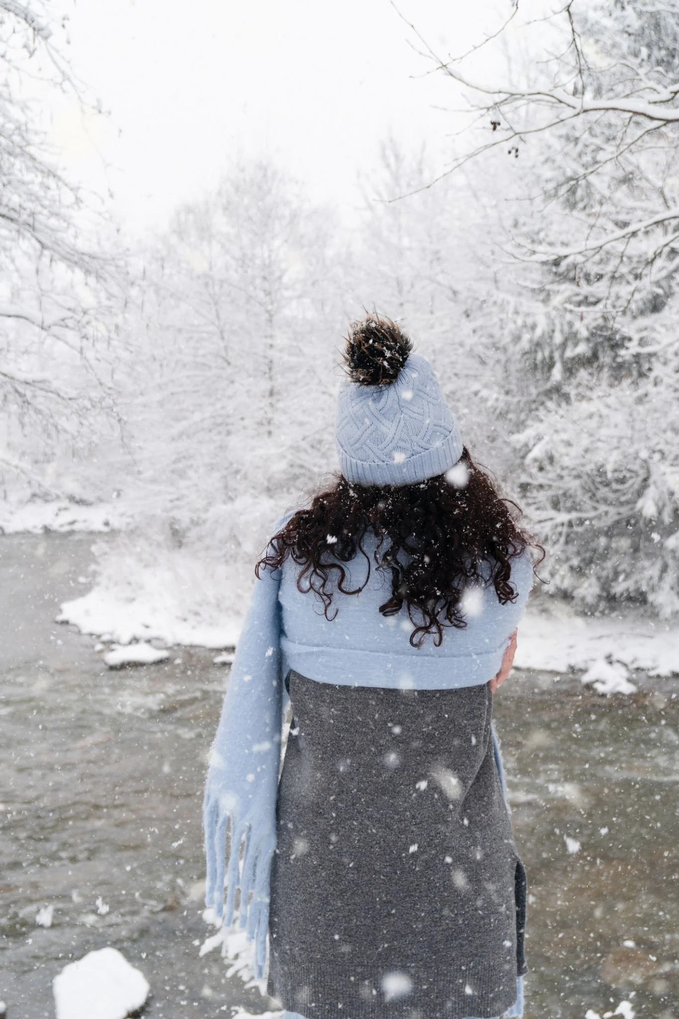 A person with curly hair wearing a gray coat, light blue scarf, and matching blue knit hat with a pom-pom, standing in a snowy landscape facing a partially frozen river surrounded by snow-covered trees.