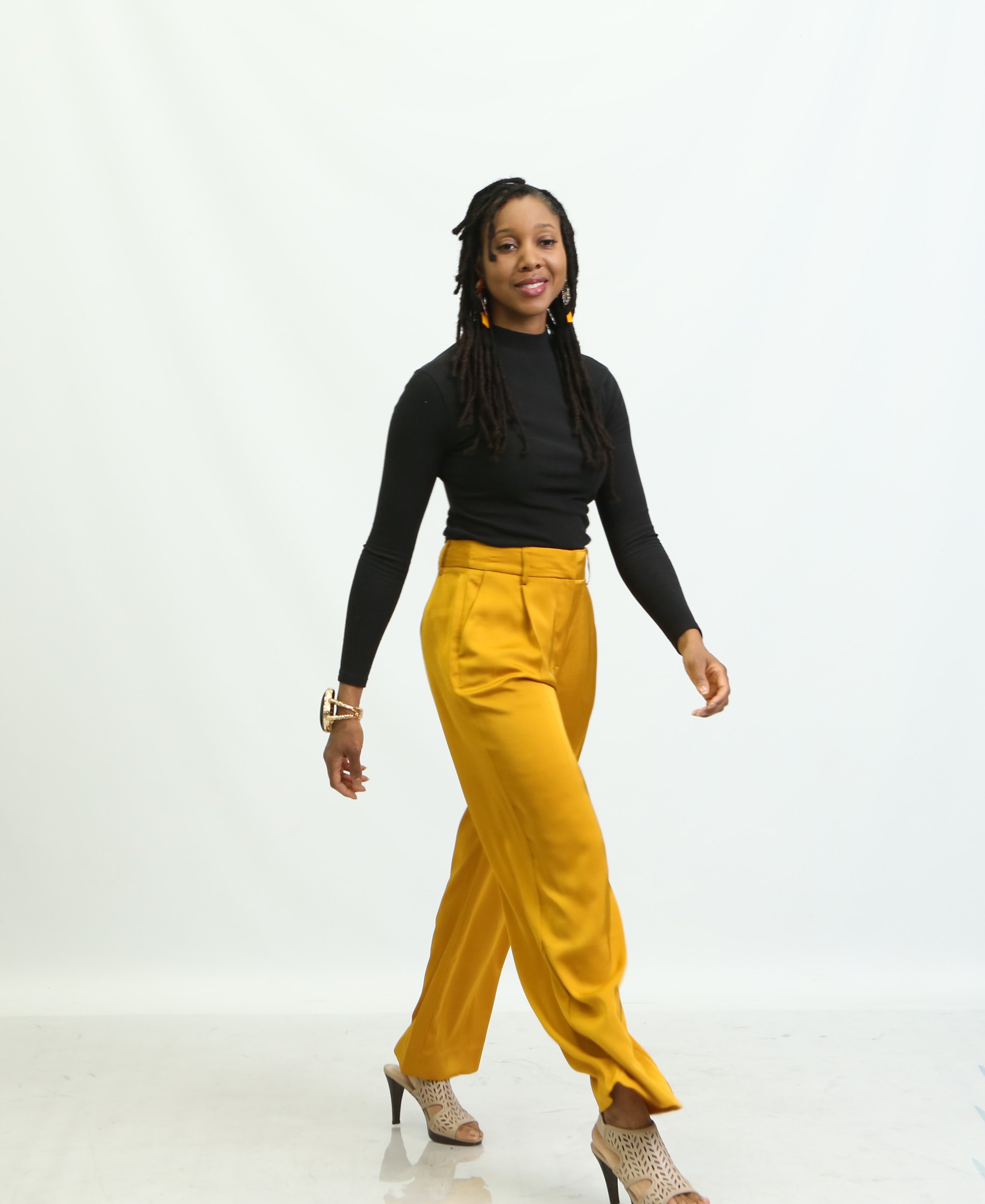 Woman walking in a studio with a white background, wearing a black long-sleeve top, yellow high-waisted pants, and high-heeled shoes.