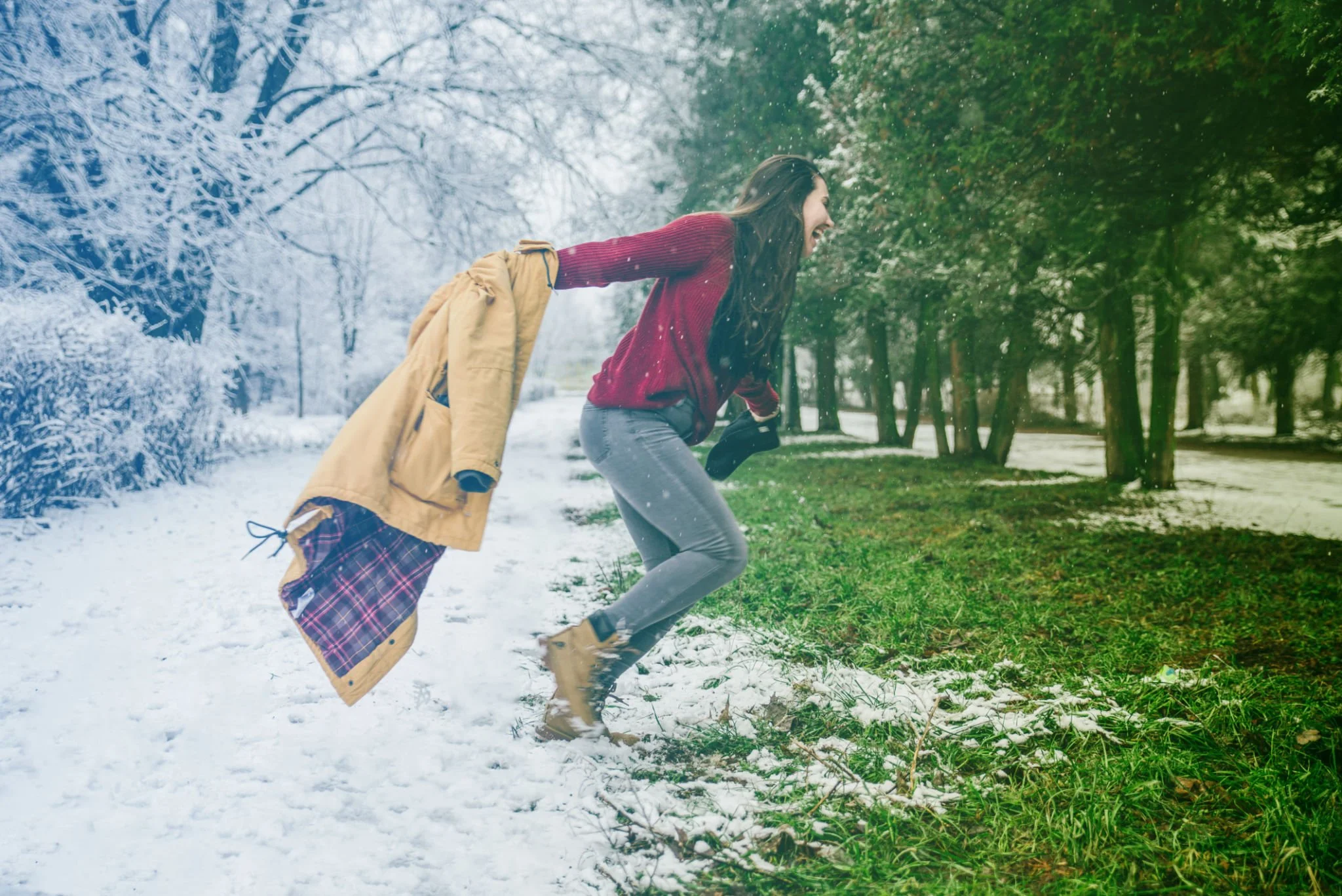 A woman with long brown hair in a red sweater and gray jeans is joyfully jumping on the border between a winter snowy area and a green grassy area, with trees in the background.