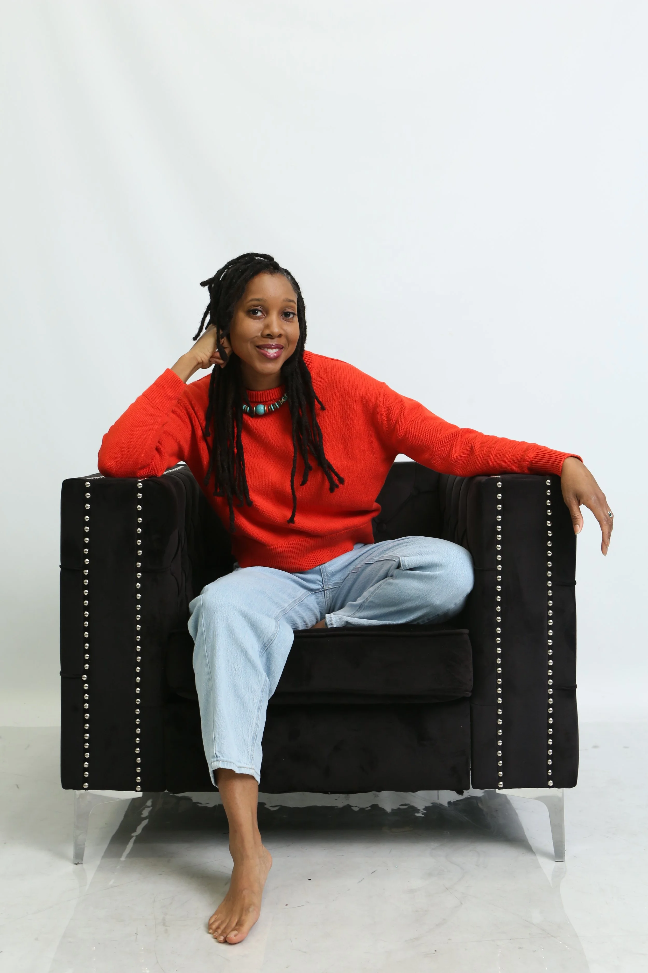 Woman sitting on a black armchair with silver studs, wearing a red sweater and light blue jeans, smiling, with one hand resting on her head and the other on the armrest, in a white room.