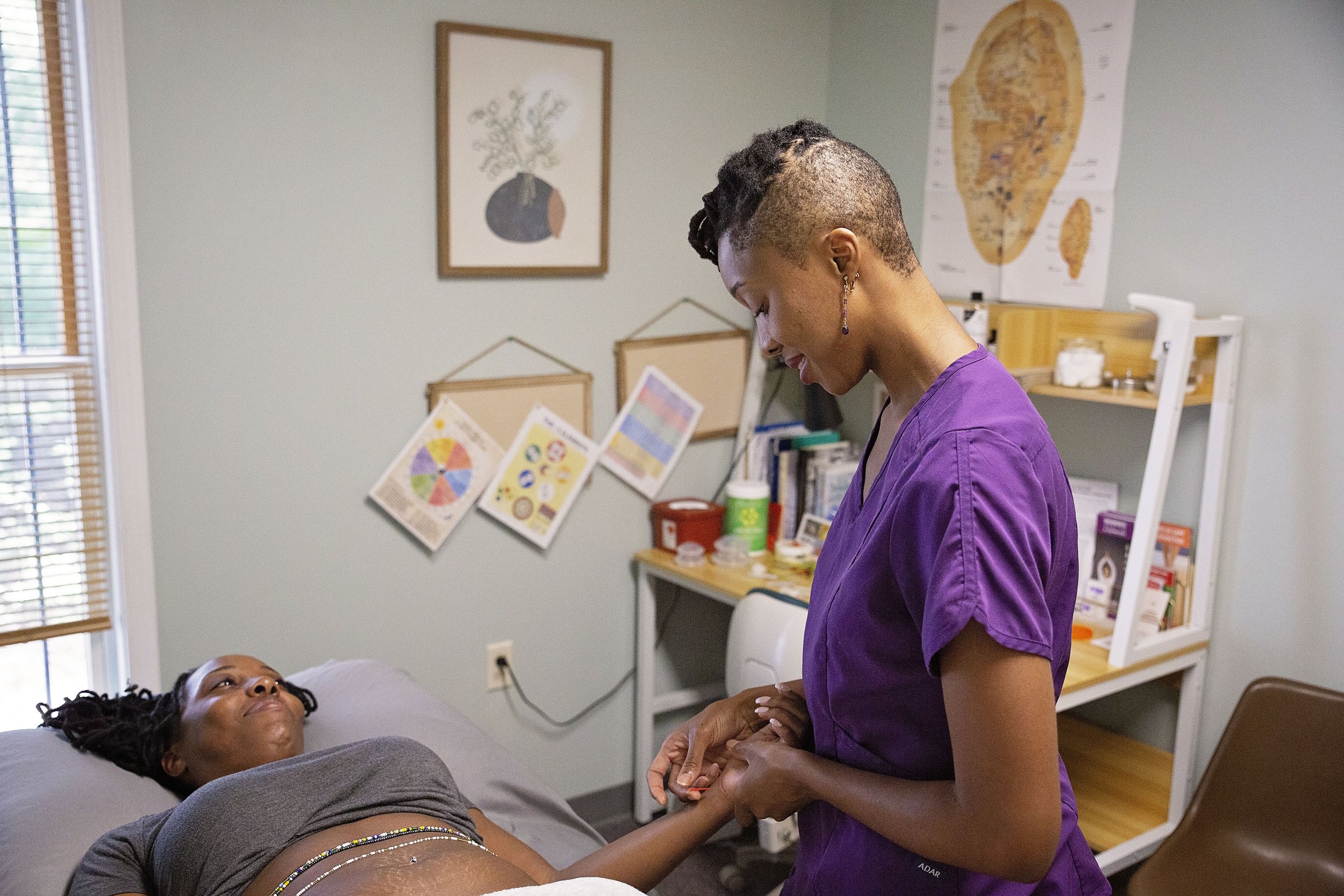 A nurse in purple scrubs holding the hand of a patient lying in a hospital bed, looking at the patient affectionately in a medical office.