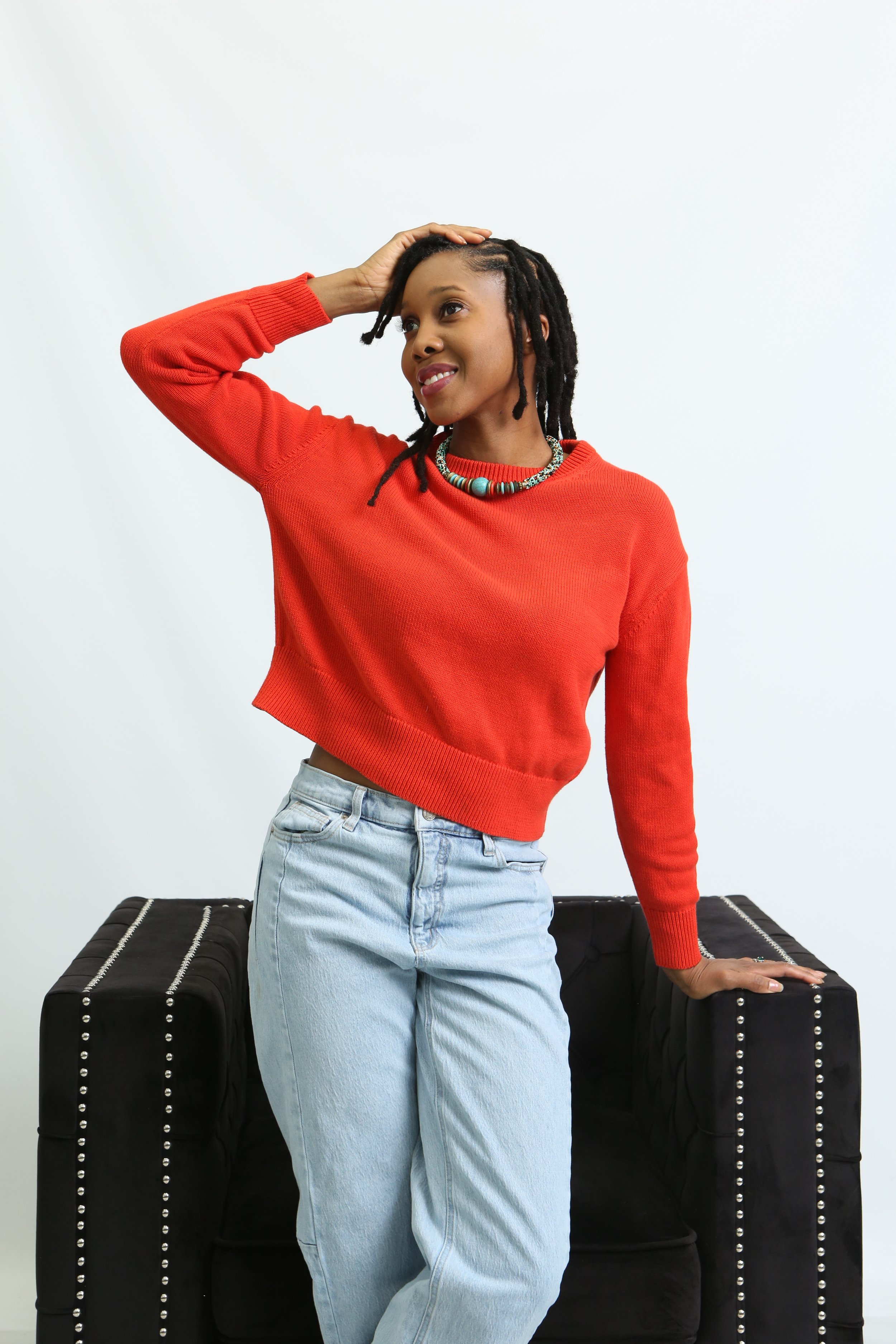 A young woman with dreadlocks in a red sweater and light blue jeans poses with one hand on her head and the other resting on a black velvet sofa with silver nailhead trim against a white background.