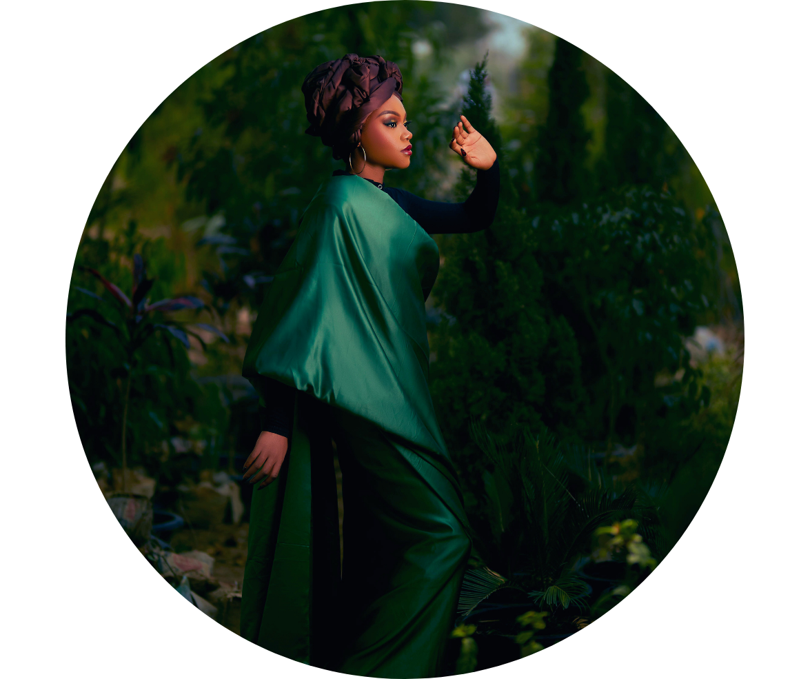 A woman in a green satin dress and black long sleeve shirt standing outdoors surrounded by lush greenery, with one hand raised and wearing a headwrap.