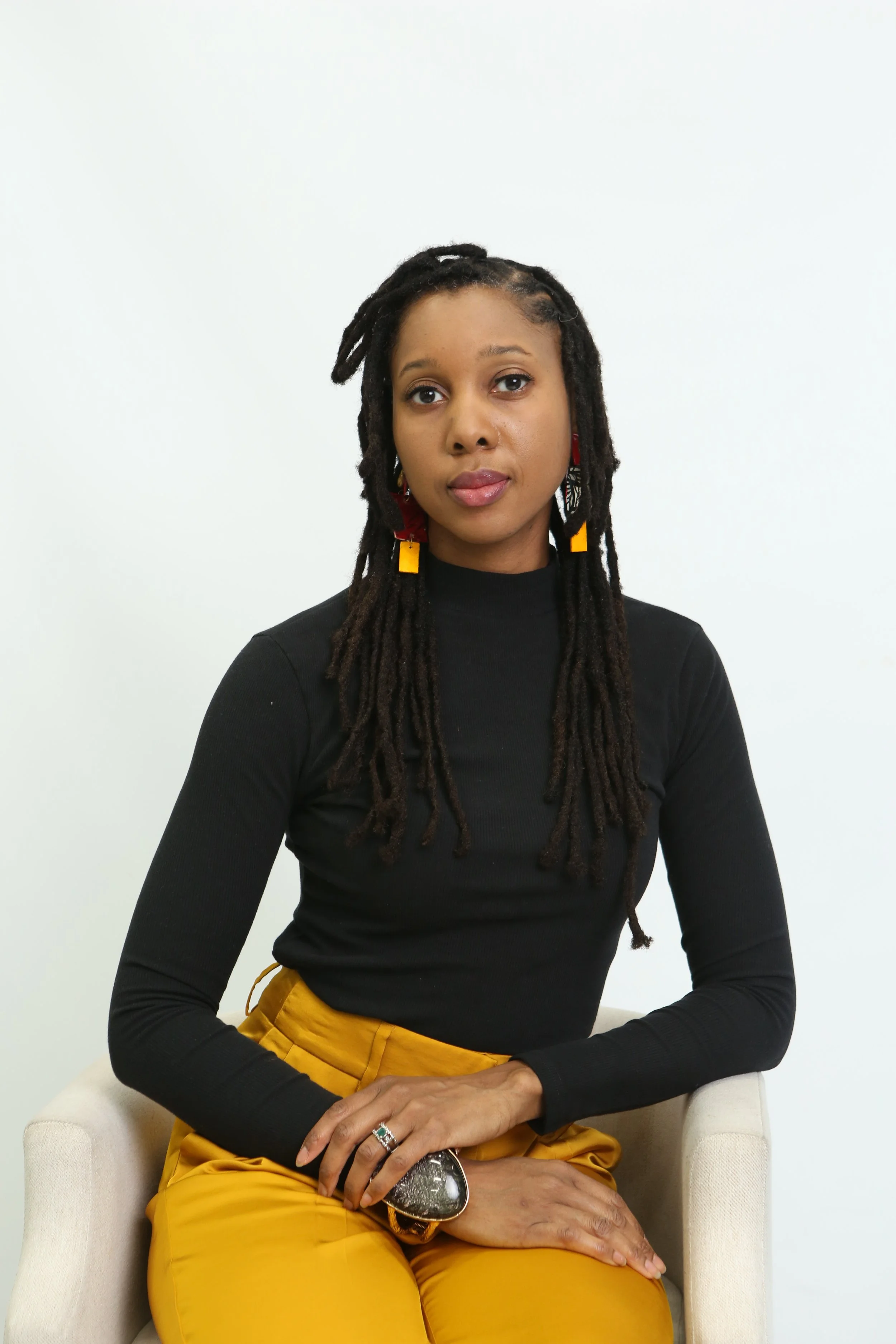 A woman with long dreadlocks sitting on a beige chair against a plain white background.
