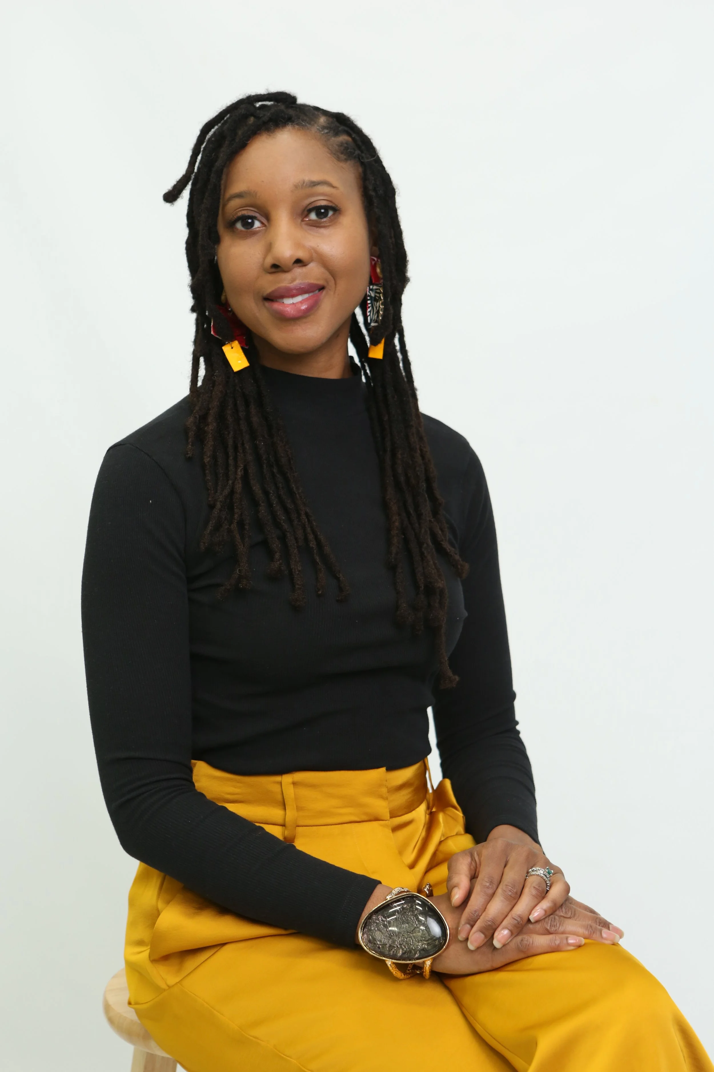 A woman with long dark dreadlocks styled with a headband, wearing a black long-sleeve shirt and mustard yellow pants, sitting on a wooden stool against a plain white background, smiling softly and looking at the camera.