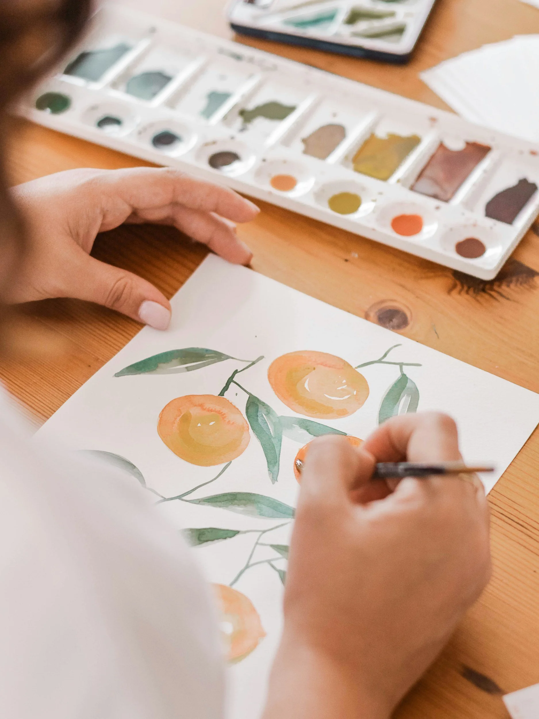 Person painting a watercolor of oranges and green leaves on paper with a watercolor palette in the background.