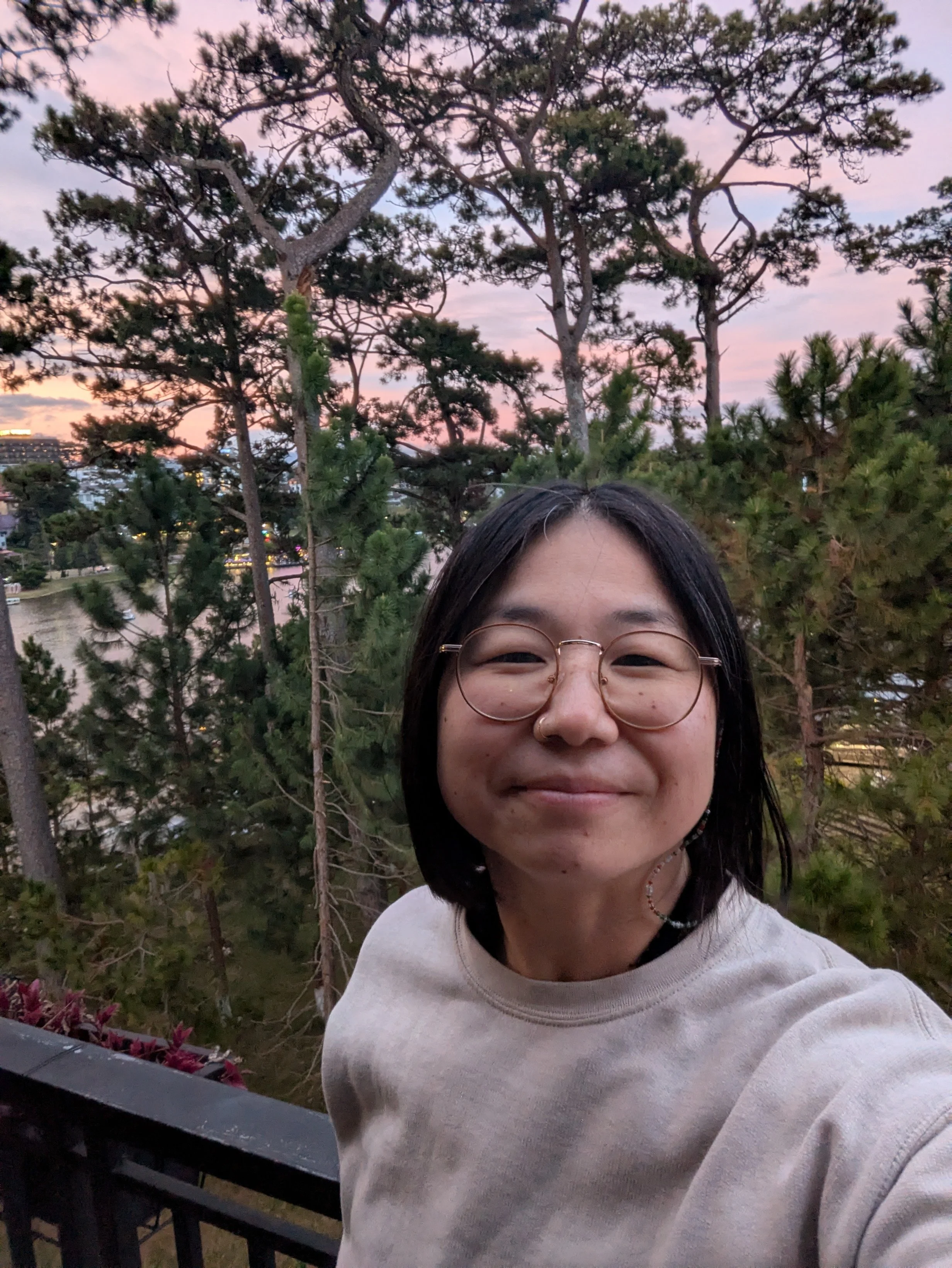 A young woman with glasses and dark hair smiling outdoors during sunset, standing in front of a backdrop of trees and a cityscape.