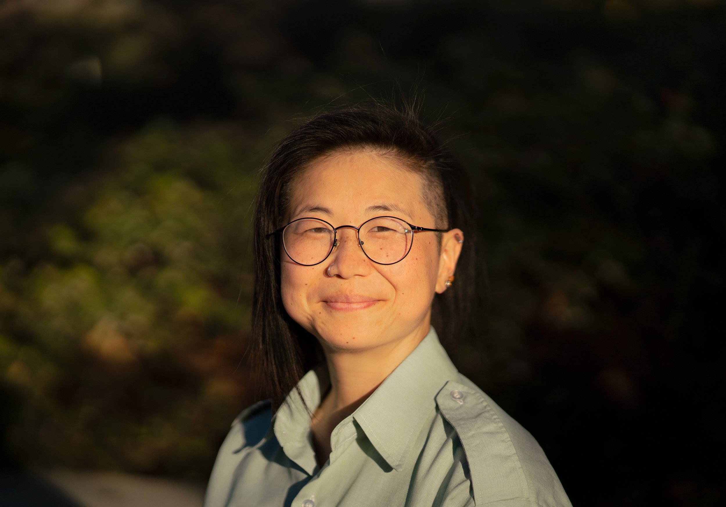 Close-up of a person with glasses, wearing a light green shirt, smiling, outdoors with dark blurred background.