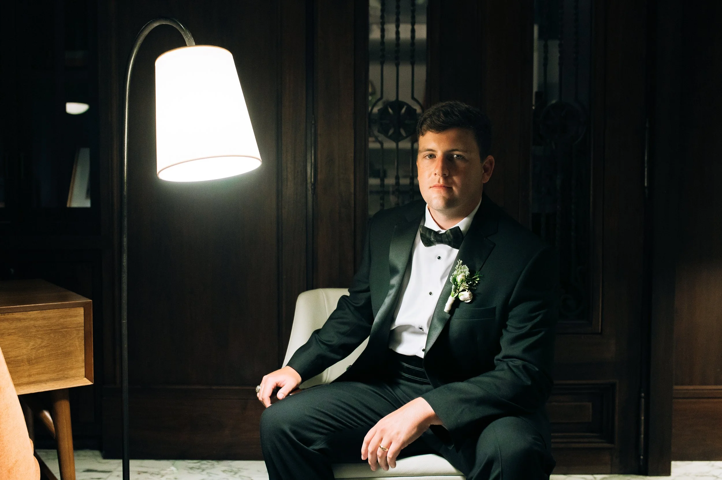 A groom in a black tuxedo with a white shirt and bow tie, sitting on a white chair in a dimly lit room, illuminated by a tall floor lamp.  Savannah wedding photography.   Wedding Photography by Sowing Clover Photography.