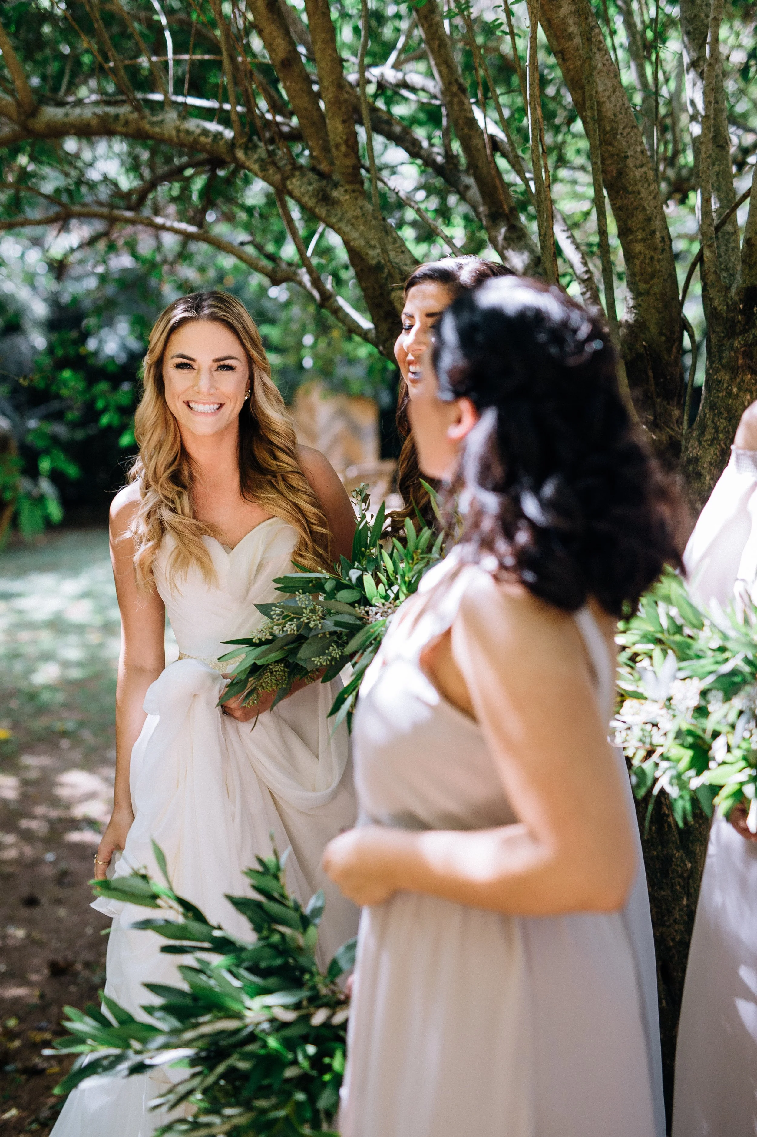 Women in elegant dresses holding bouquets, standing outdoors under a tree, smiling and chatting.  Mint Springs Farm in Tennessee. Wedding photography by Sowing Clover Photography.