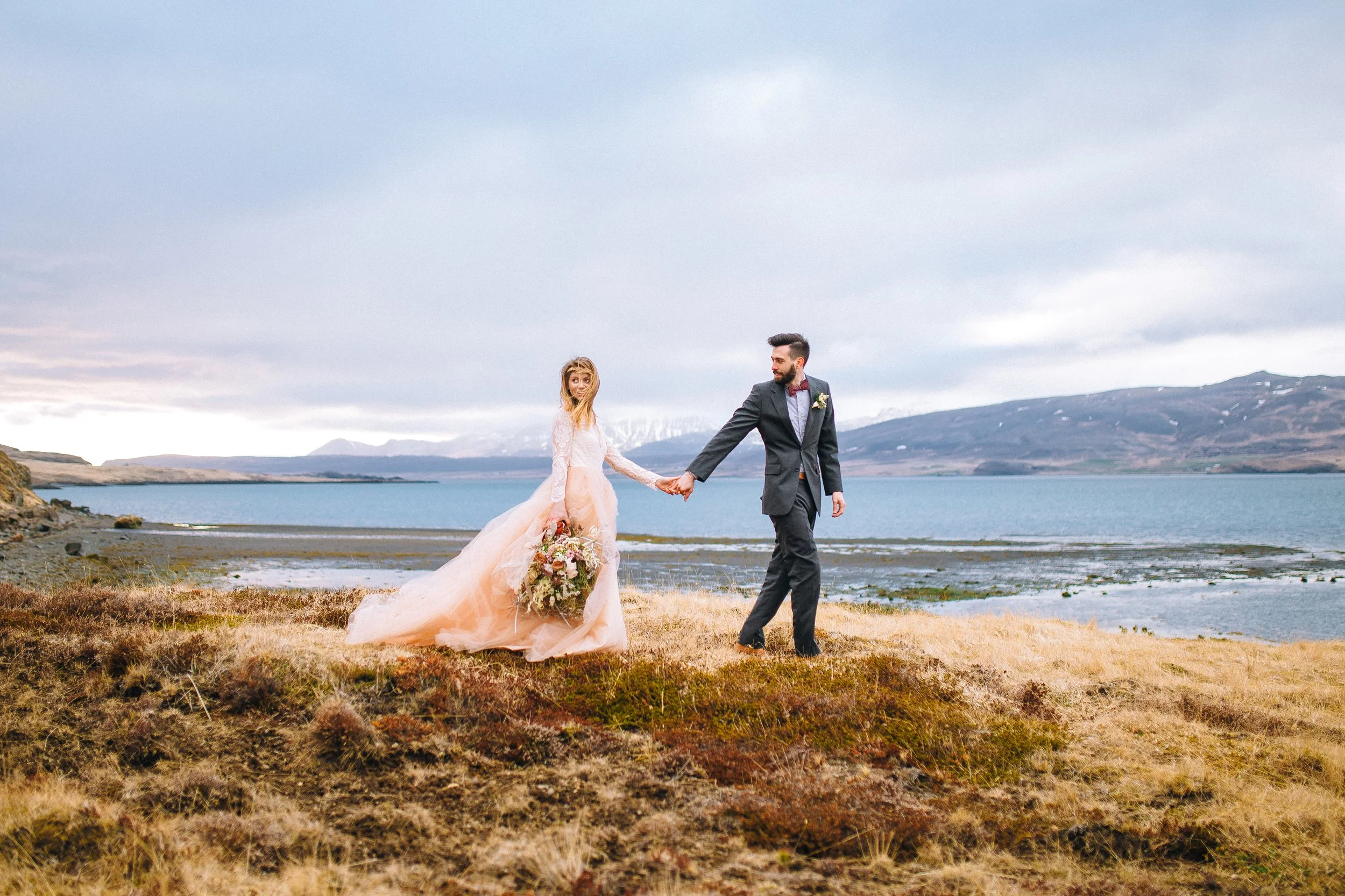 Bride in a peach wedding gown holding a bouquet and groom in a black suit holding hands, standing on grassy terrain near a body of water with mountains in the background in Iceland. Wedding Photography by Sowing Clover Photography