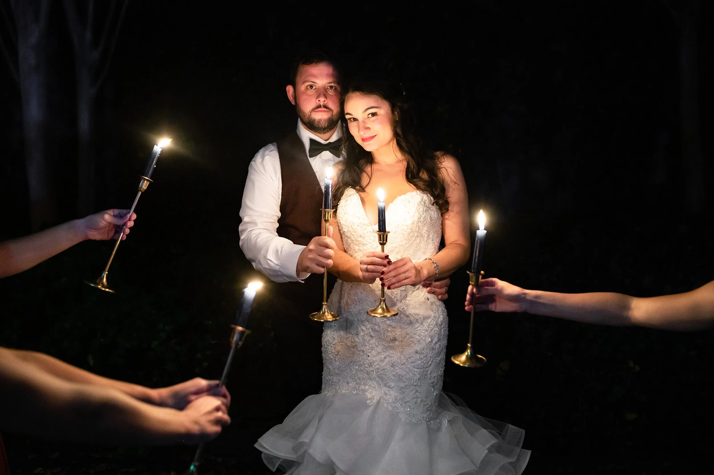 Bride and groom holding candles during a nighttime wedding photoshoot, surrounded by arm holding candles in a dark outdoor setting. Wedding Photography by Sowing Clover Photography. In Atlanta